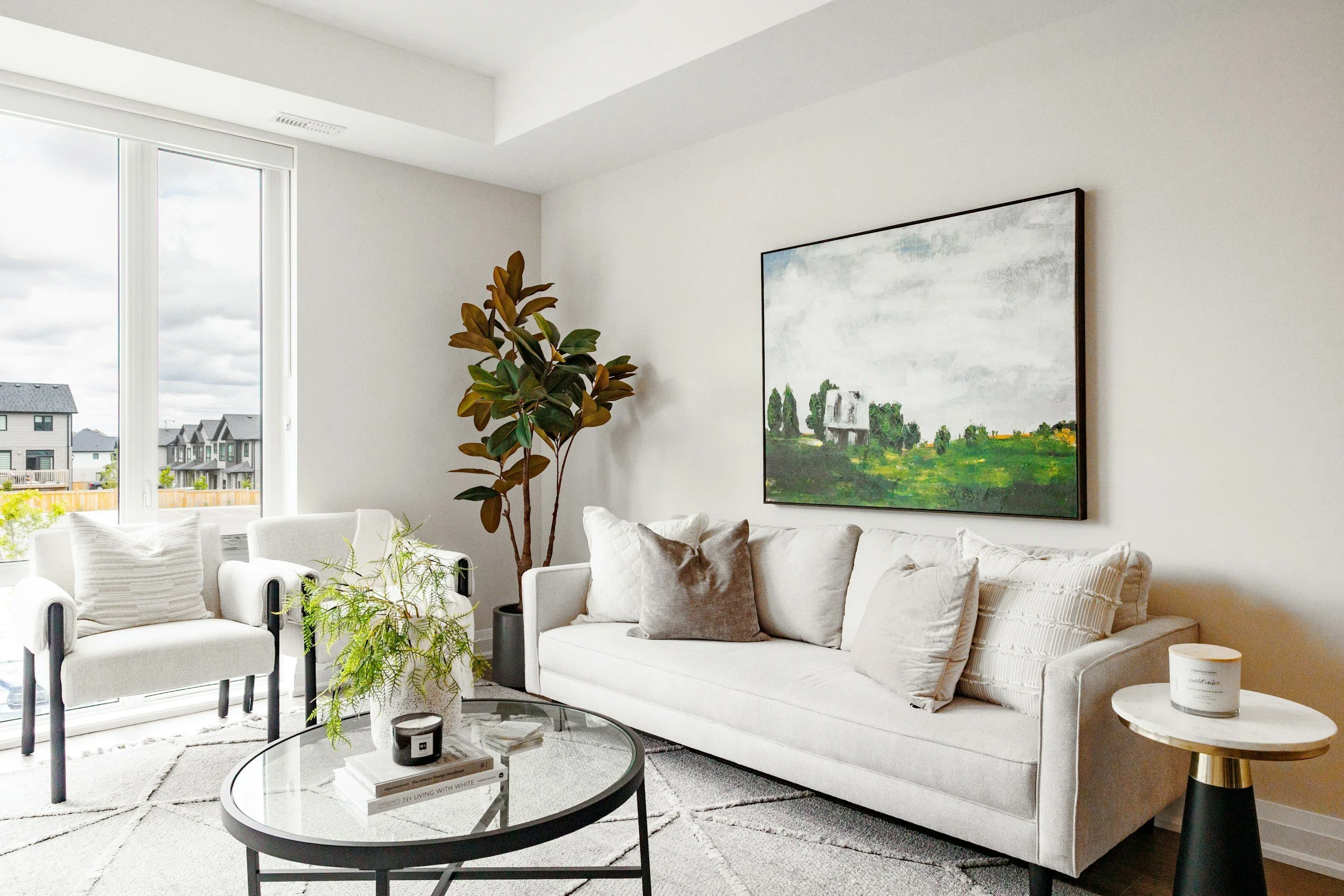 Modern living room with white sofa, gray pillows, a glass coffee table, potted plants, artwork on the wall, and large windows with a view of neighboring houses.