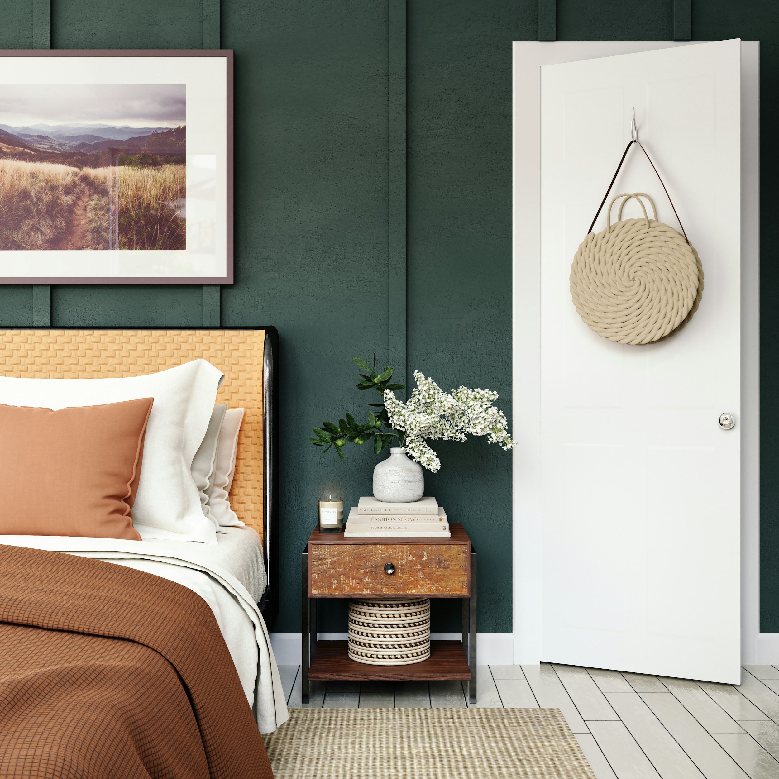 A bedroom corner with a green textured wall, a framed landscape photograph, a bed with a woven headboard and neutral-colored pillows, a wooden nightstand with a white vase of white flowers, a small candle, and a stack of books. A white door with a beige round woven bag hanging on it is to the right.