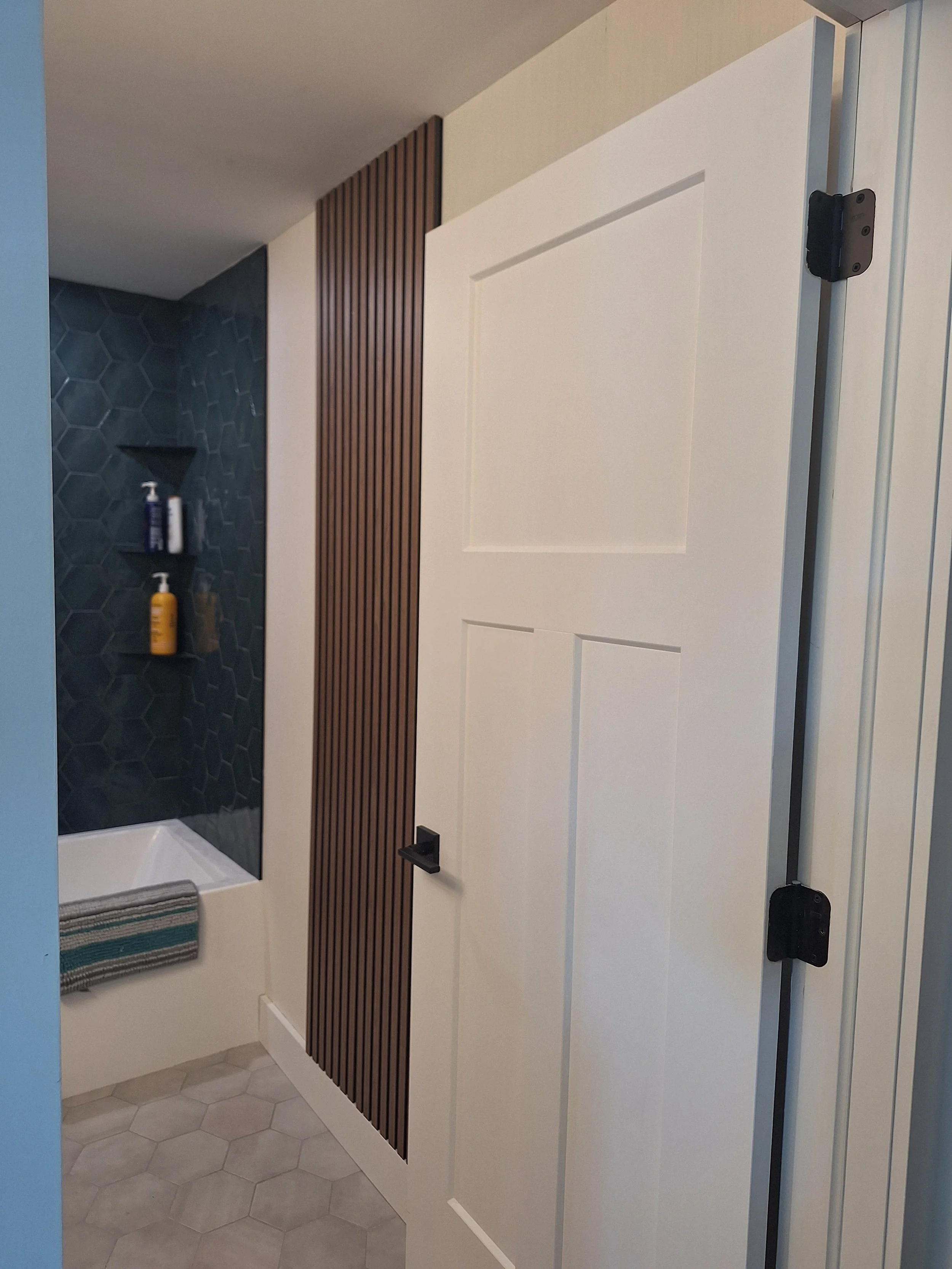 A modern bathroom with a white door, a blue hexagon tile shower wall with built-in shelves holding shampoo bottles, and a wooden slatted panel beside the door. The floor has hexagon tiles.