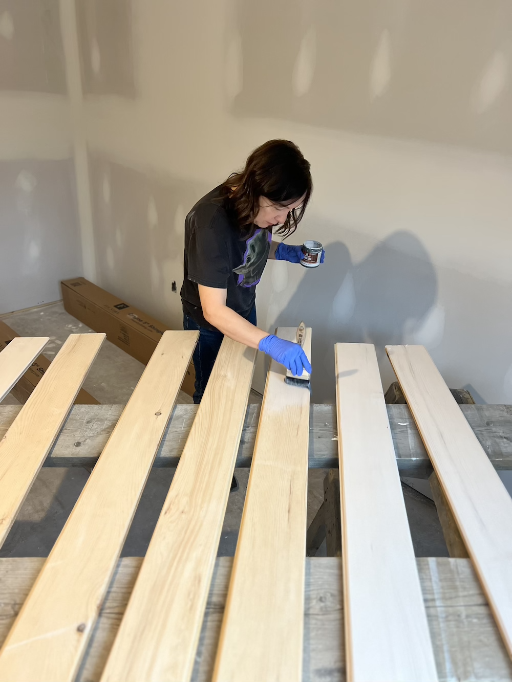 A woman in a black t-shirt and blue gloves is applying a stain or finish to wooden slats with a brush, working on a construction or woodworking project in a room with unfinished walls.