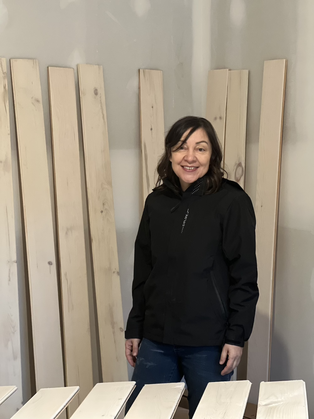 A woman with shoulder-length dark hair, smiling, wearing a black jacket, standing in front of wooden planks in a room under construction or renovation.