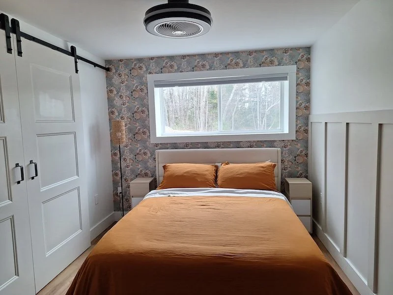 A neatly made bed with brown bedding and pillows in a bedroom with a floral accent wall, a window with a view of trees, white nightstands, a tall floor lamp, white closet doors, and a ceiling fan with a light.