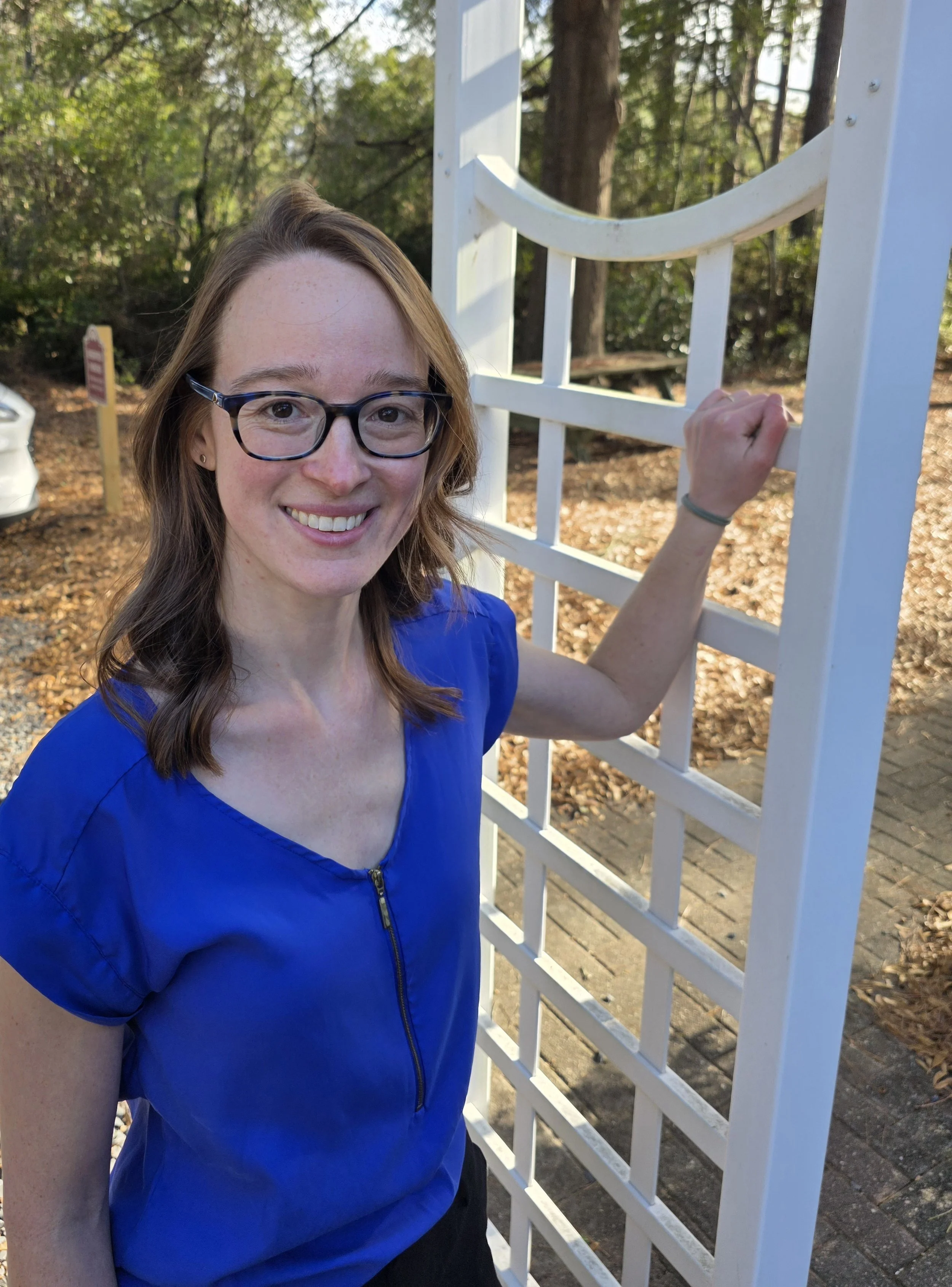 A smiling woman with glasses and wavy brown hair, wearing a royal blue top with a zipper, standing next to a white garden trellis outdoors, surrounded by trees and fallen leaves.