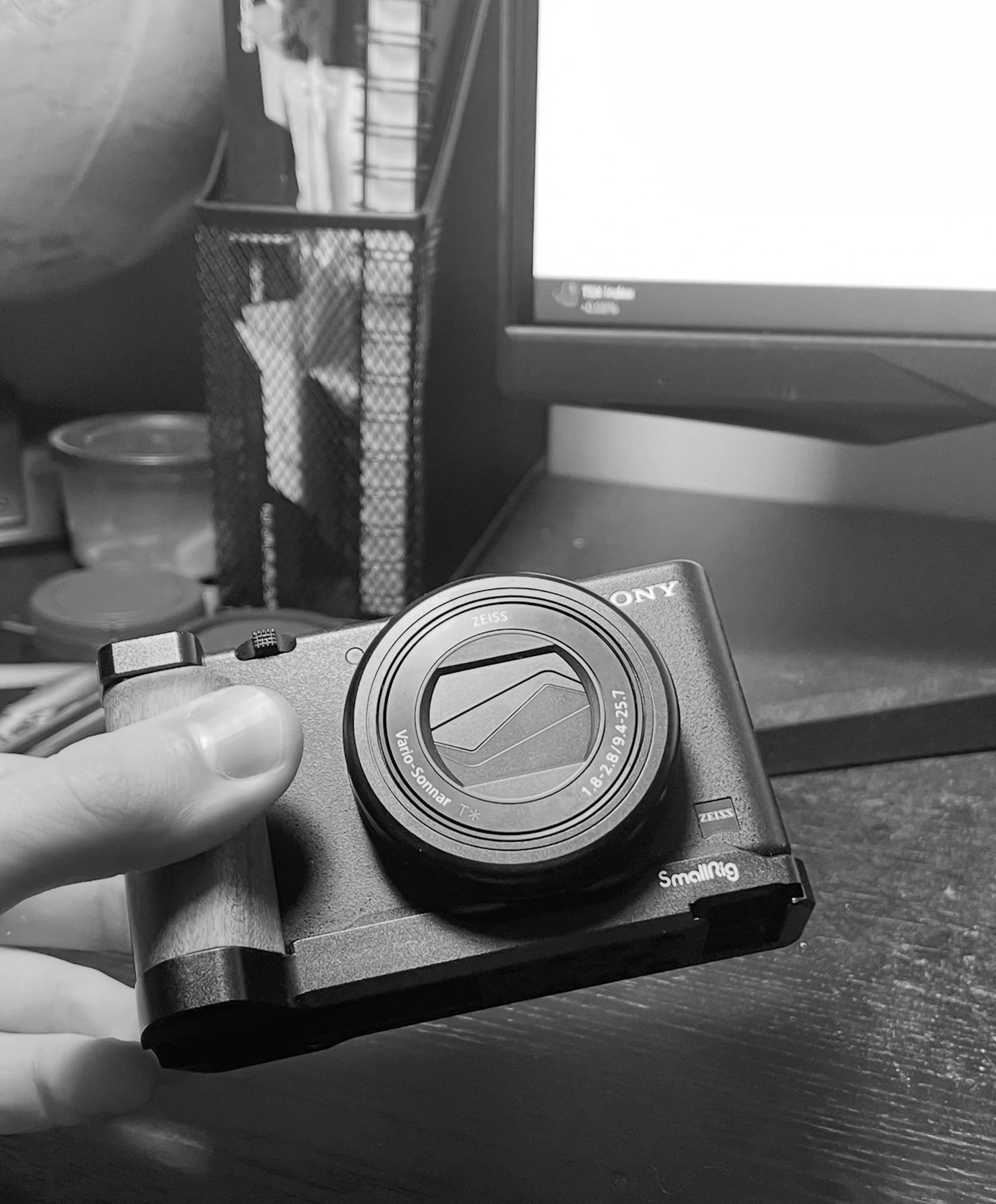 A digital camera being held in front of a computer monitor on a desk, with office supplies in the background.