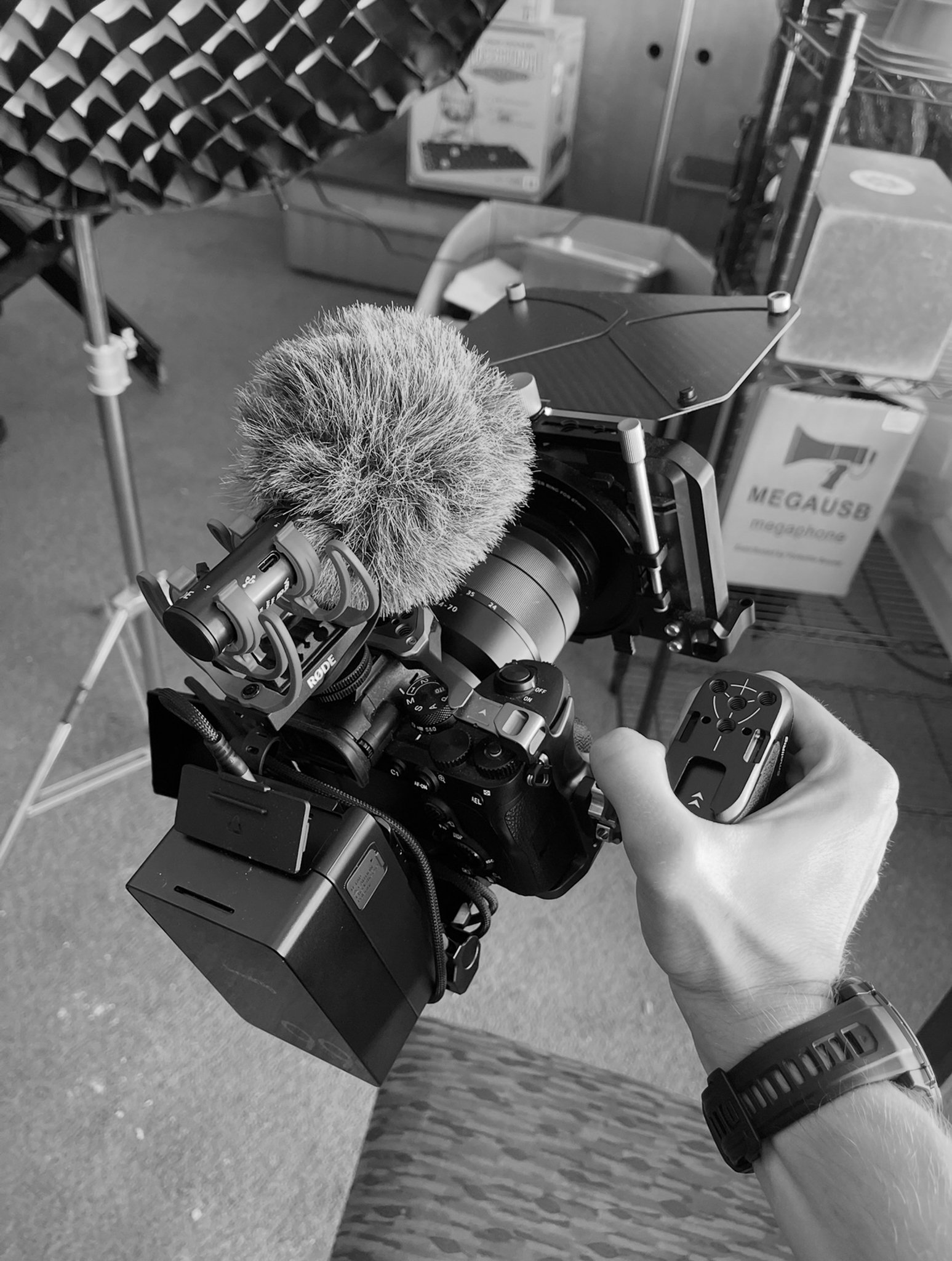 A person holding a professional camera with a microphone attached, in an indoor setting with various equipment and storage boxes in the background.
