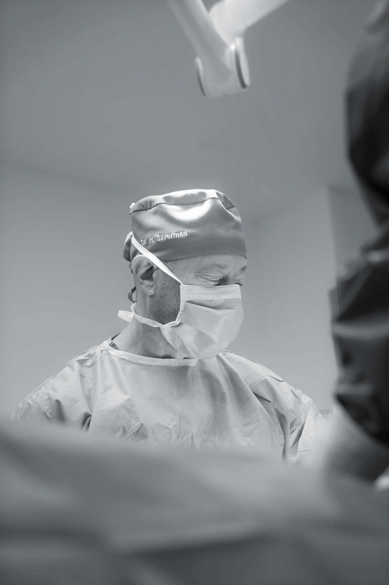 A medical professional wearing surgical scrubs, a cap with "Dr. H. Silverman" embroidered, and a face mask, appears to be performing surgery or a medical procedure in an operating room.