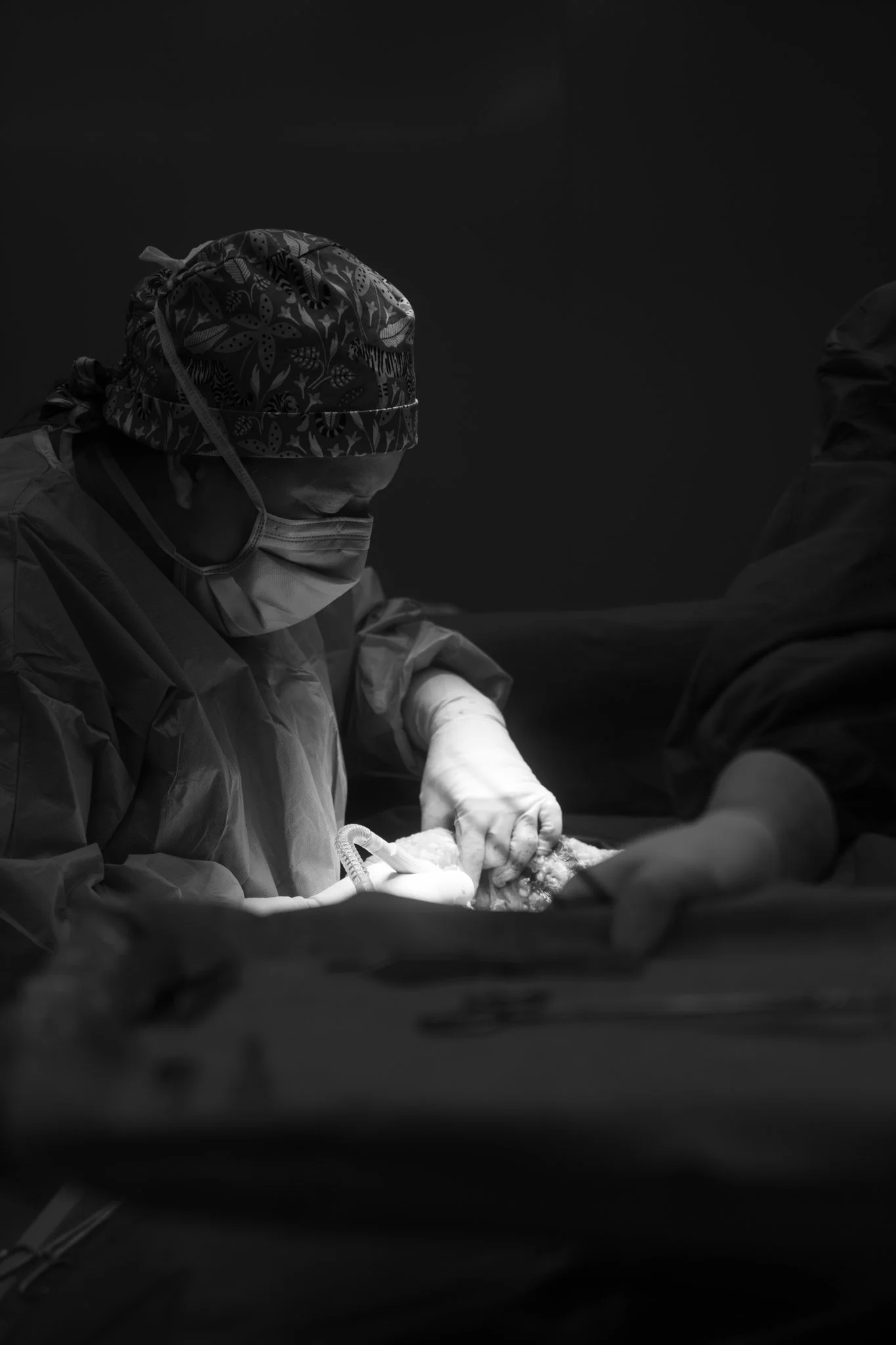 A medical professional in scrubs and a face mask performing surgery in a dark operating room.