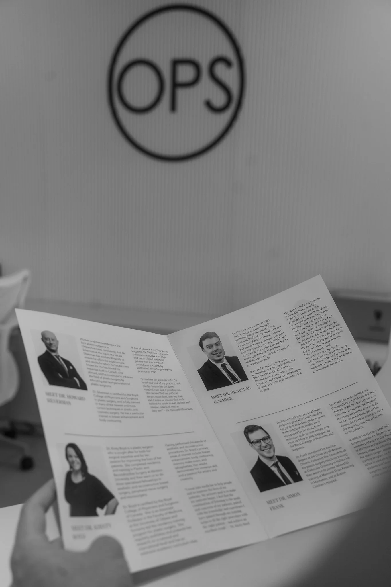 Black and white photo of a hand holding a booklet featuring profiles of three people, including photos and descriptions, in front of a wall with a circular OPS logo.