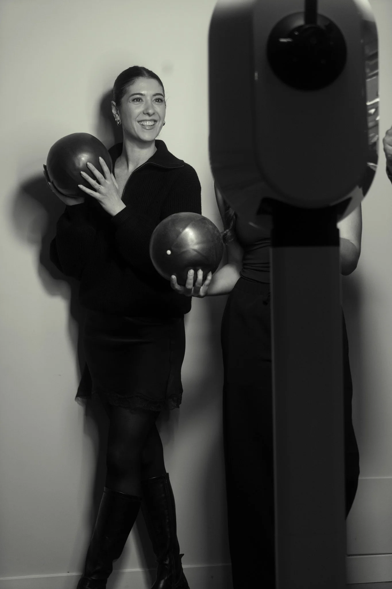 A woman smiling and holding two kettlebells in front of a plain wall, with a exercise machine partially visible.
