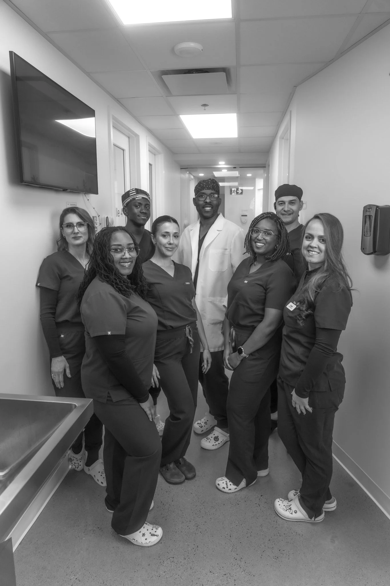 A group of healthcare professionals, including seven women and one man, standing in a hospital corridor, smiling at the camera. They are wearing scrubs and medical footwear.