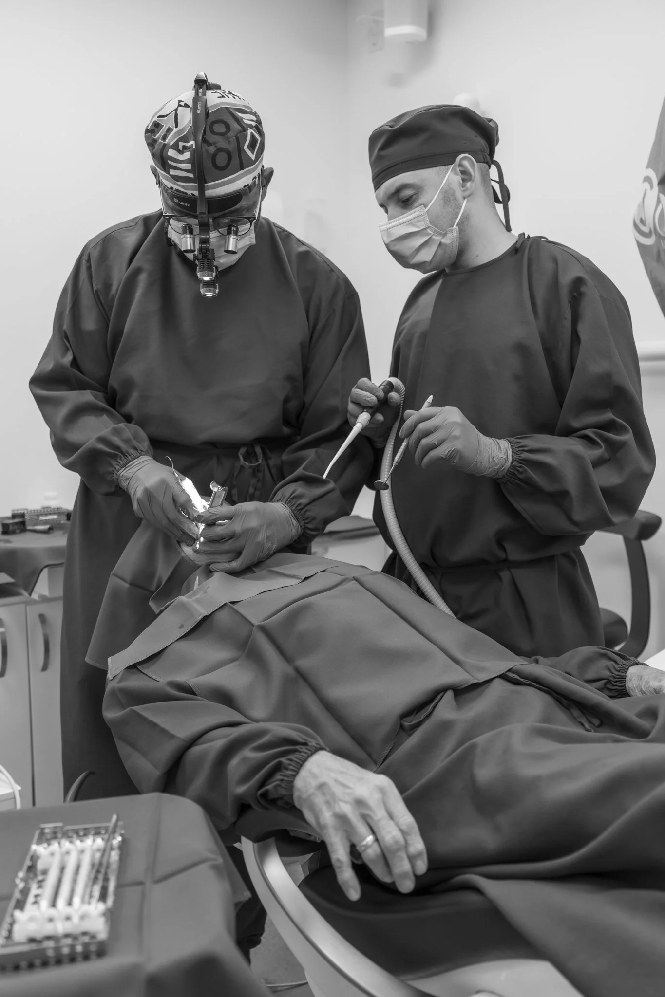 Two surgeons performing surgery on a patient in an operating room, both wearing surgical masks, gowns, gloves, and one with a surgical headlamp.