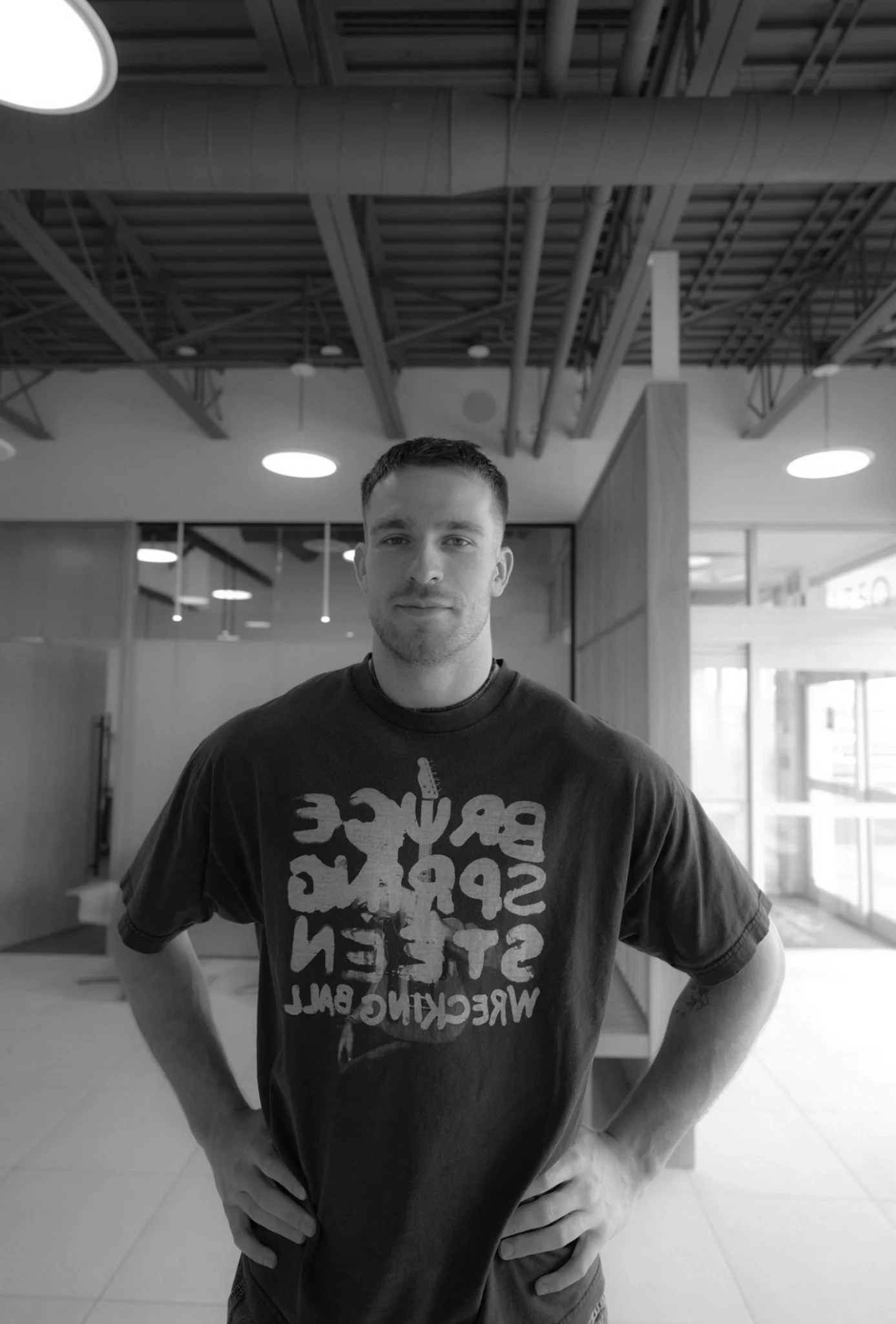 A young man with short dark hair and a slight beard standing indoors with hands on hips, smiling slightly. He is wearing a T-shirt with a graphic design and text, in a modern interior space with visible ceiling pipes, round lights, and large windows in the background.