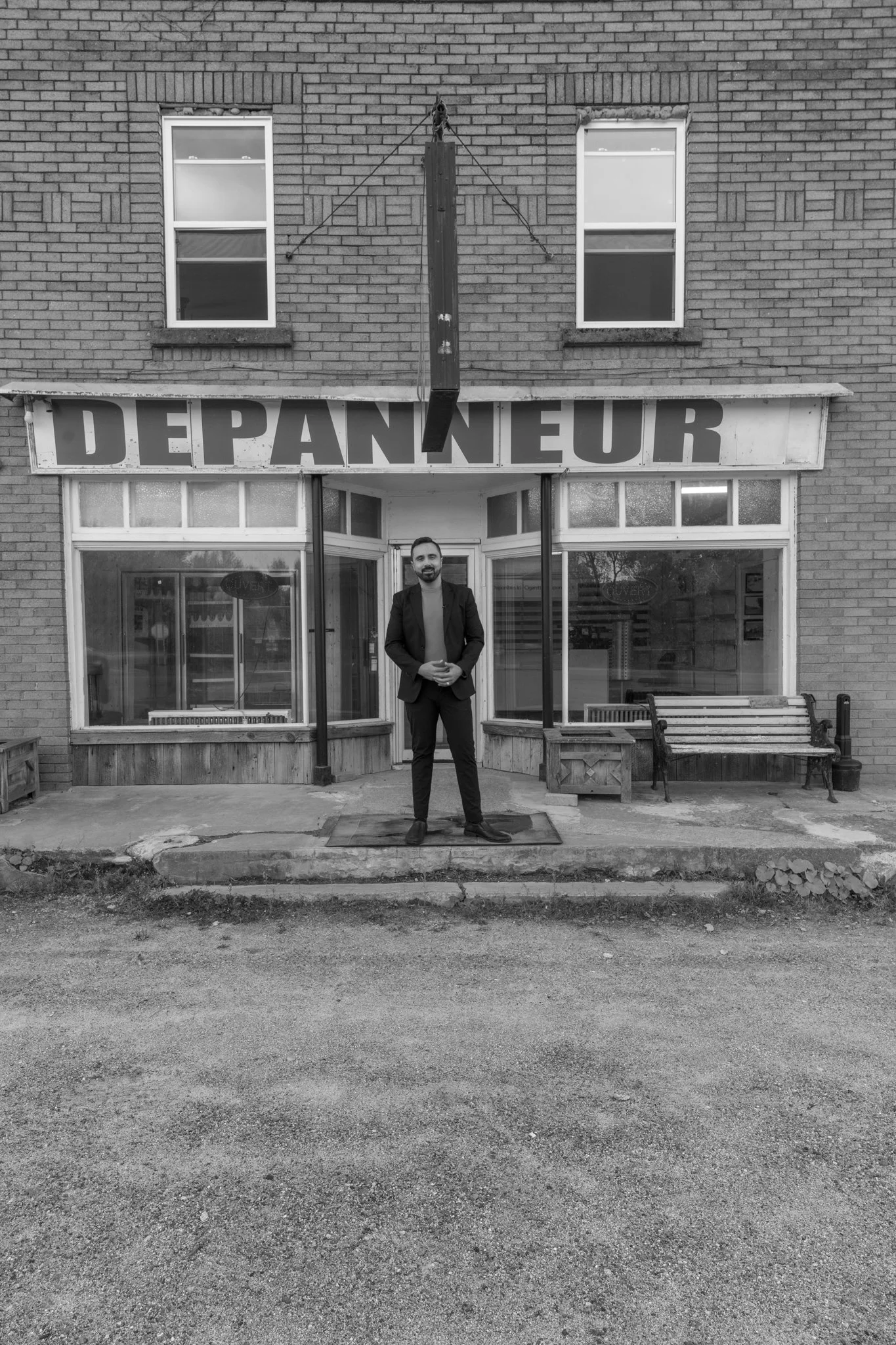 A man in a suit standing outside a vintage storefront with a sign that reads 'DEPANNNEUR'.