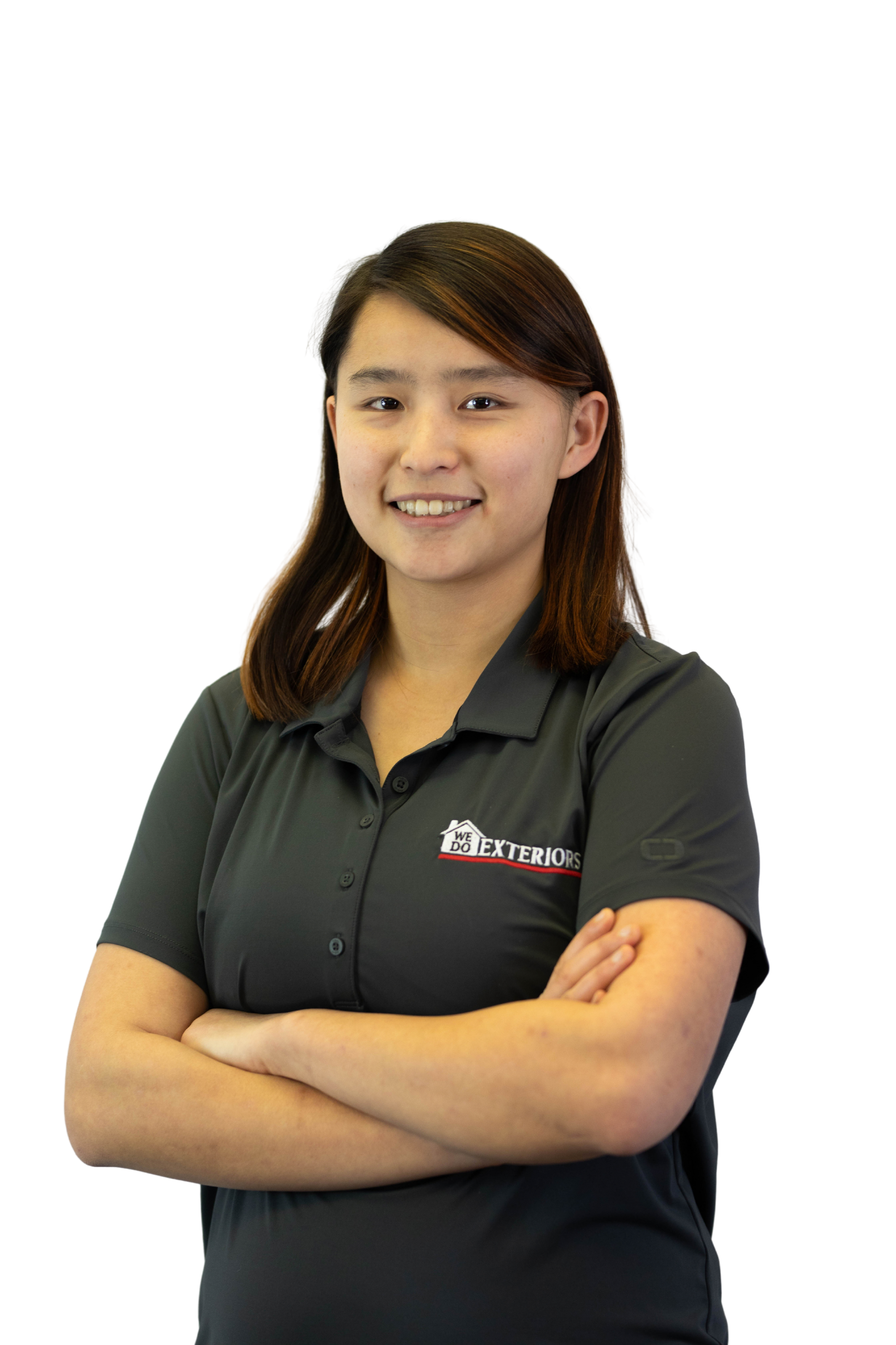 Young woman with shoulder length black and auburn hair,  smiling at the camera with her arms crossed, wearing a branded grey polo style shirt, against a white background.