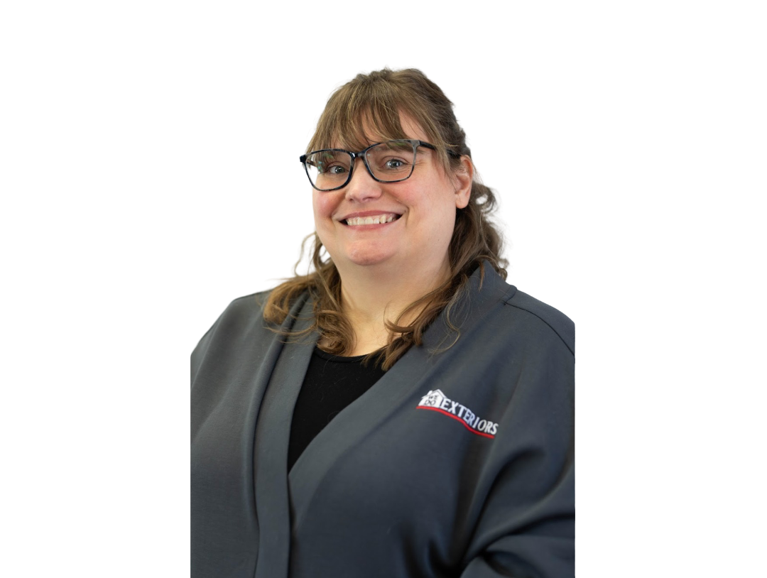 A woman with shoulder length brown hair and dark frame glasses, smiling at the camera, wearing a branded grey cardigan, against a white background.
