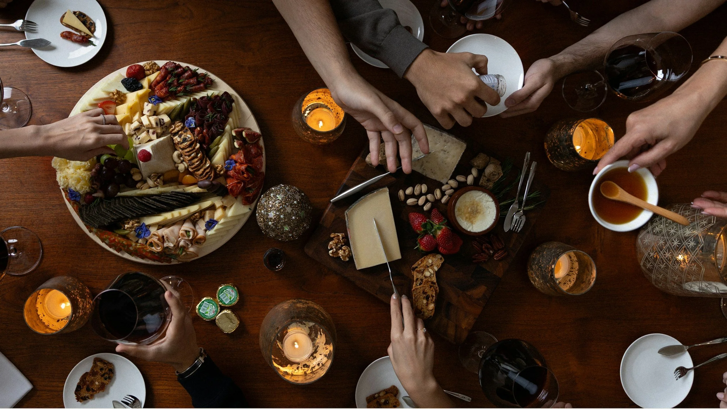 Top-down view of a dinner table with a large cheese and fruit platter, assorted cheeses, strawberries, grapes, dried fruits, nuts, and finger foods, surrounded by wine glasses, candles, and people reaching for food and drinks.