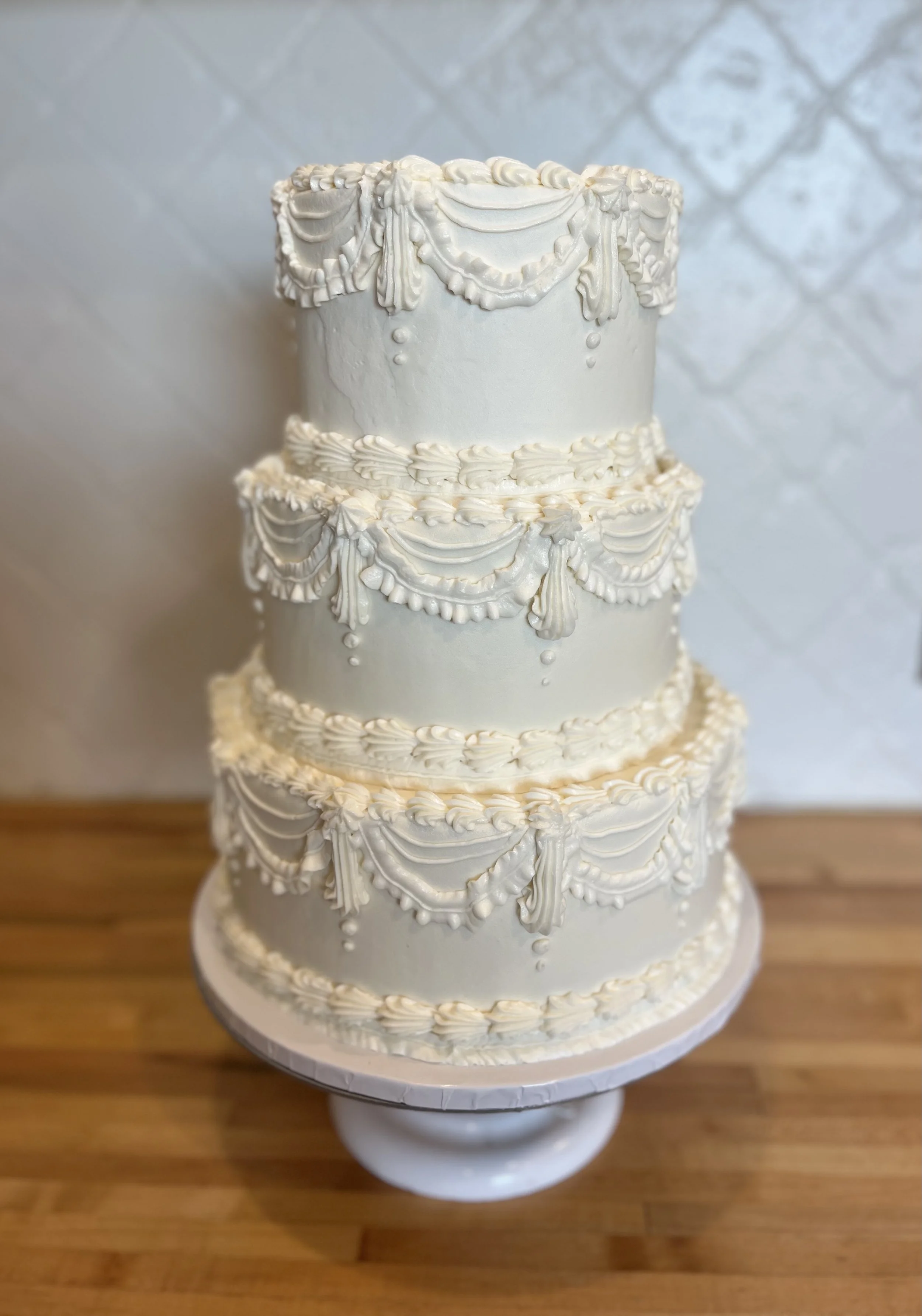 A tiered wedding cake decorated with elegant white icing, featuring intricate piped patterns and draped icing accents, placed on a white cake stand on a wooden surface.