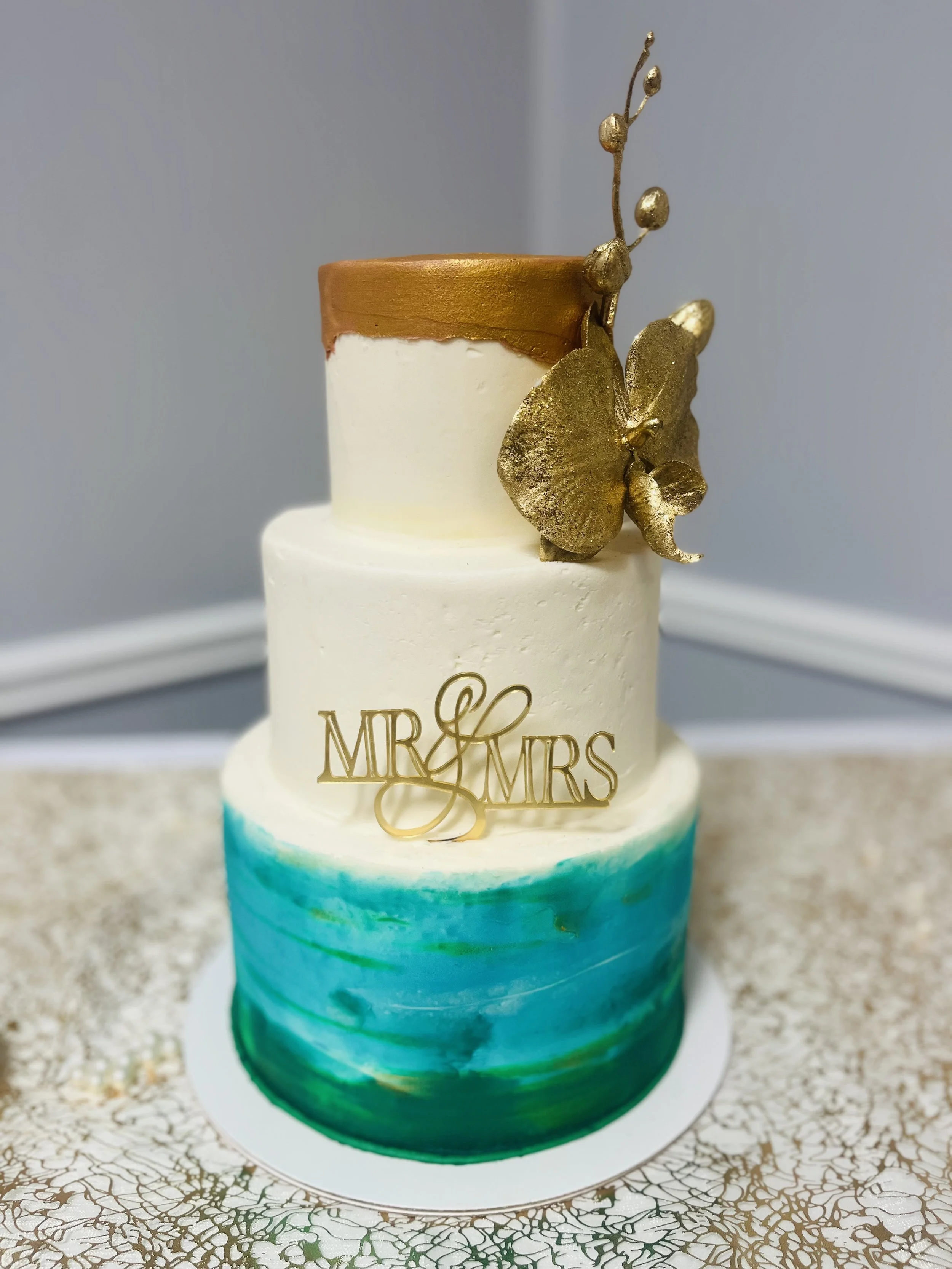A tiered cake with a watercolor ocean design at the bottom and a white upper layer, topped with a gold 'Mr & Mrs' cake topper and a decorative gold sculpture resembling a branch with leaves and berries.
