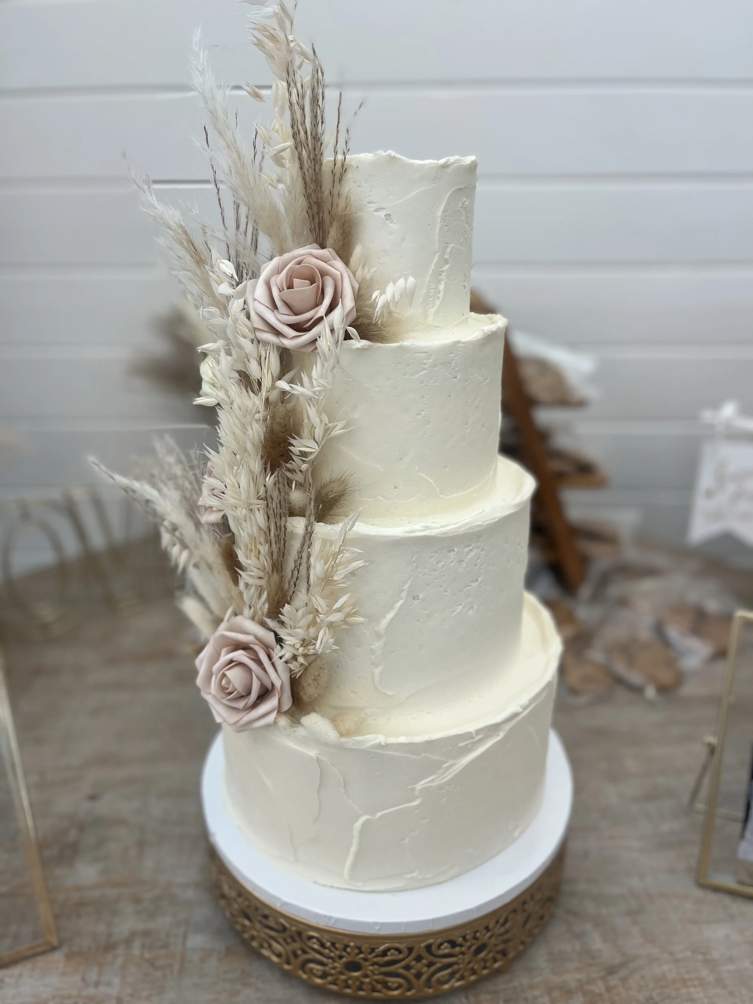 Three-tier white wedding cake decorated with light pink roses and dried beige and white flowers, with a white background.