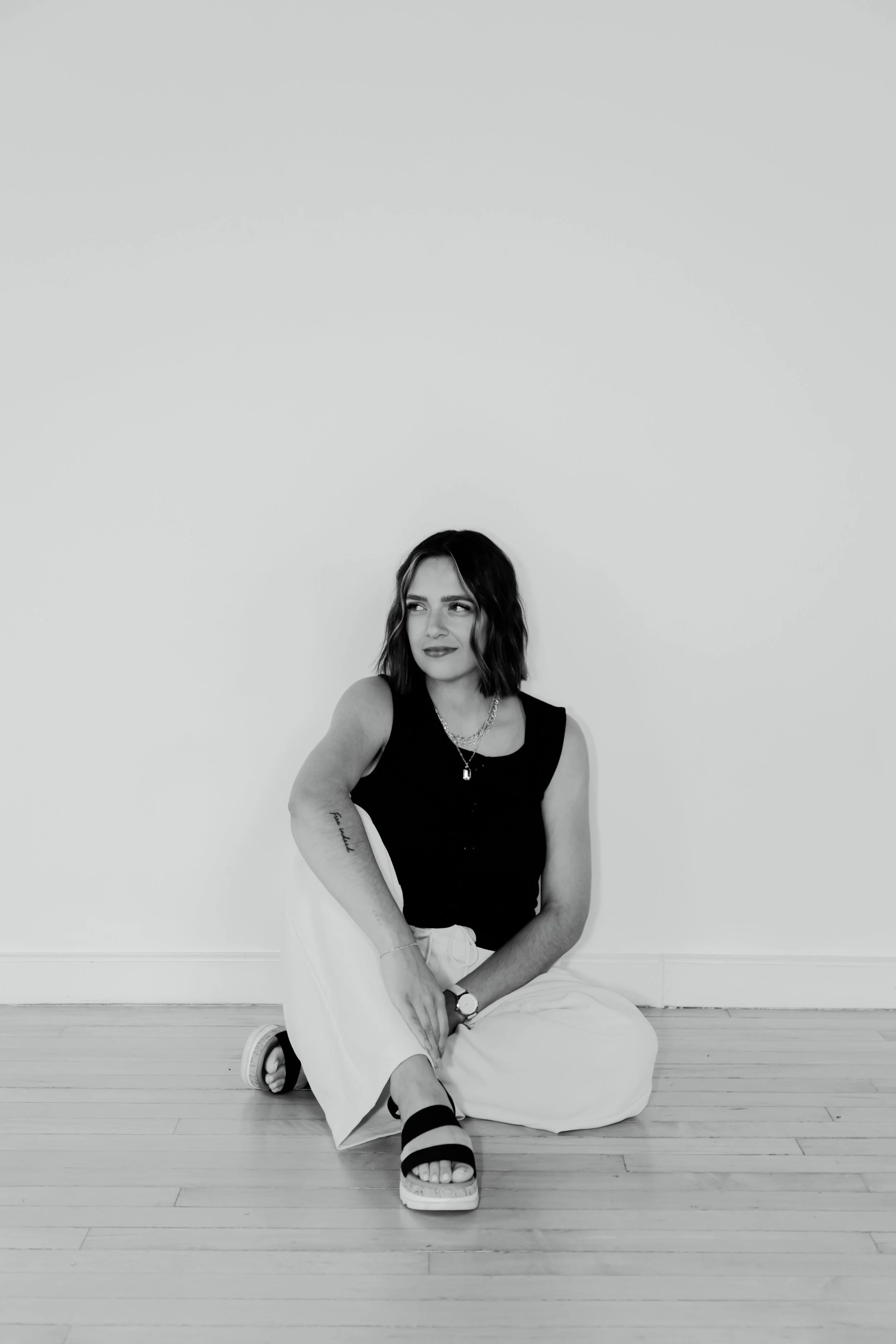 Black-and-white photo of a young woman sitting on the floor against a plain wall. She is wearing a sleeveless black top, white pants, and black sandals, with jewelry including a necklace and a watch. She has short wavy hair and is looking slightly to the side with a thoughtful expression.