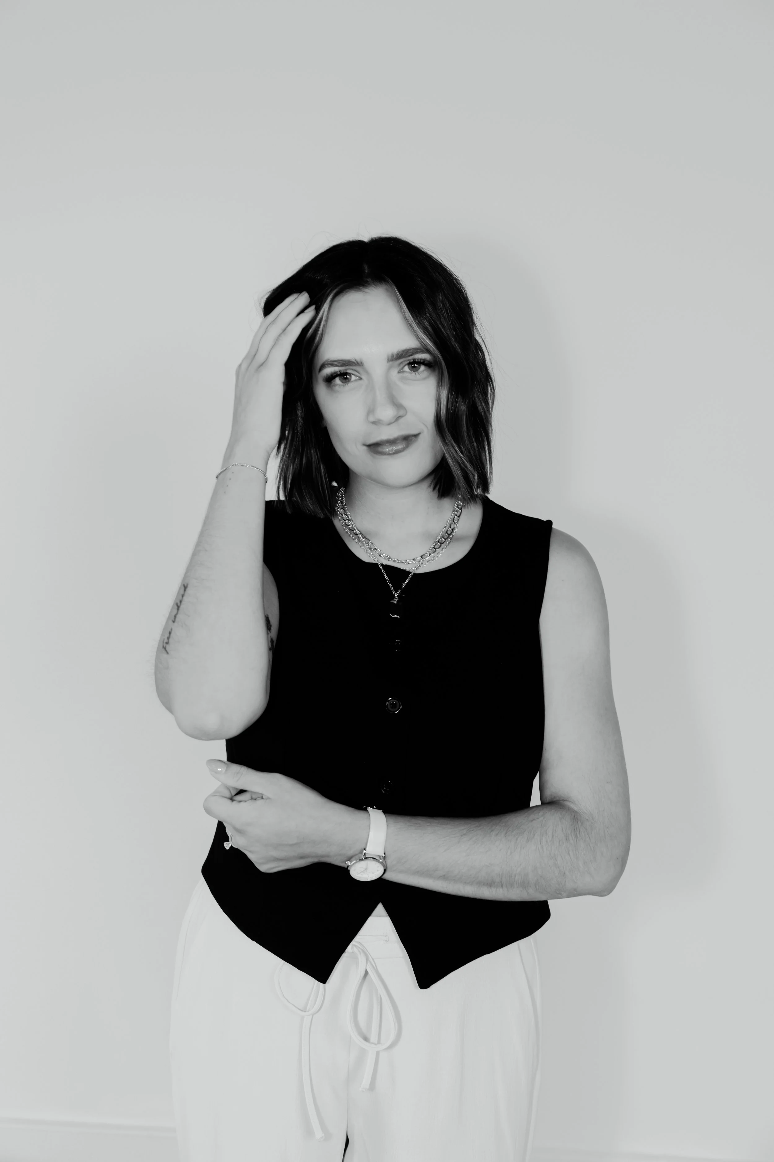 Black and white portrait of a woman with shoulder-length dark hair, wearing a sleeveless black top, white pants, a watch, layered necklaces, and bracelets, standing against a plain light background.