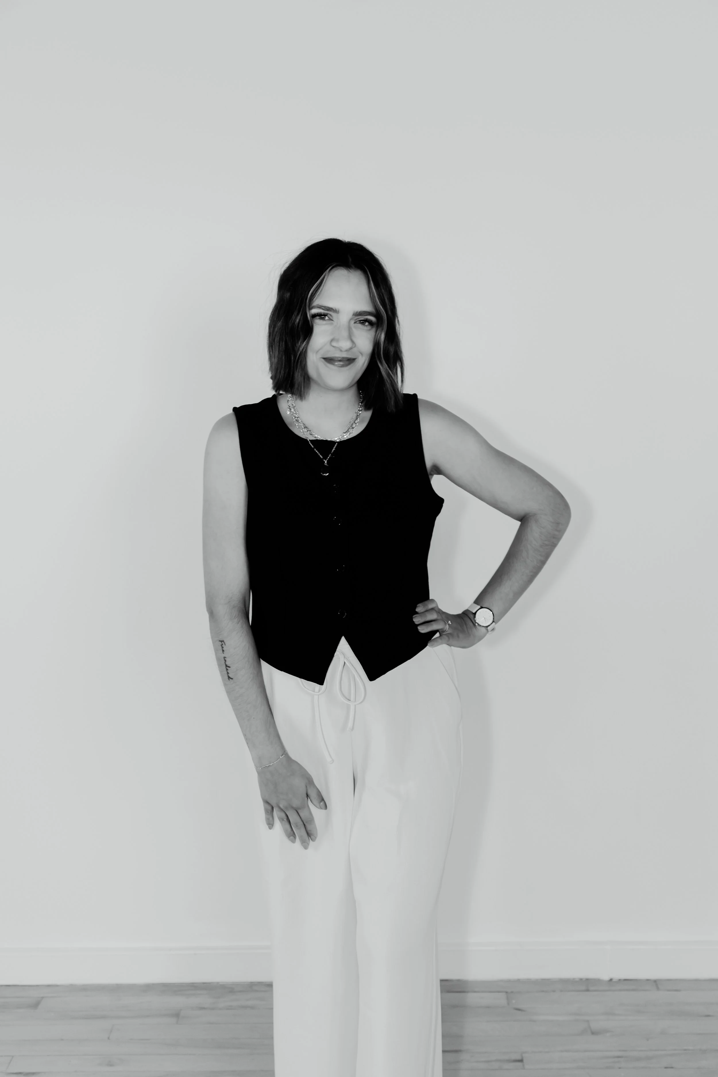 A woman with shoulder-length dark hair wearing a black sleeveless top, white pants, and jewelry, standing against a plain wall and smiling at the camera.