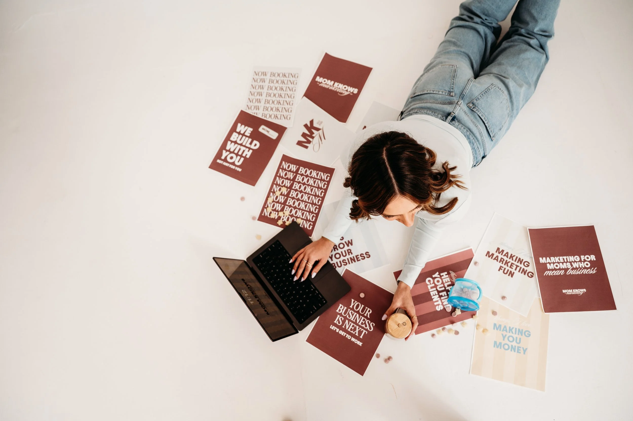 A woman lying on her stomach on a white floor, surrounded by marketing and business related pamphlets and brochures, working on a laptop.