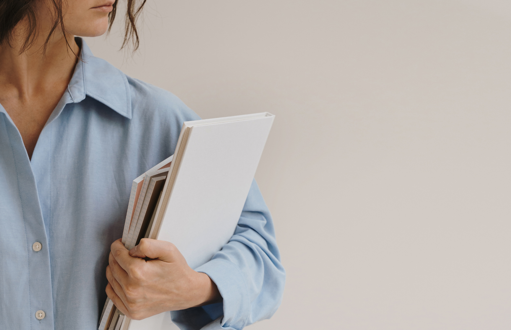 Person wearing a blue shirt holding a stack of notebooks or folders against a plain beige background.