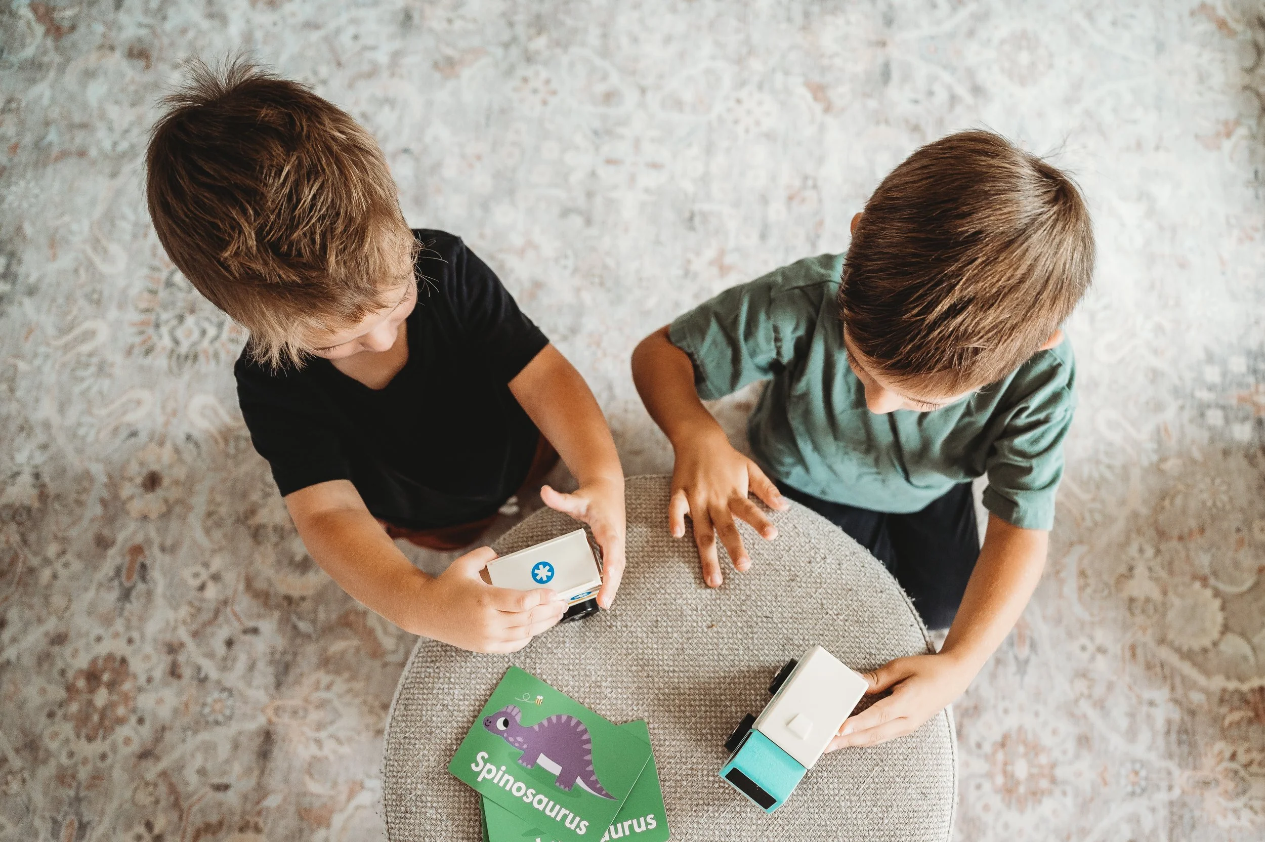 Two children playing with educational toys on a round table, one holding a sensor device, with a dinosaur flashcard labeled 'Spinosaurus' on the table.