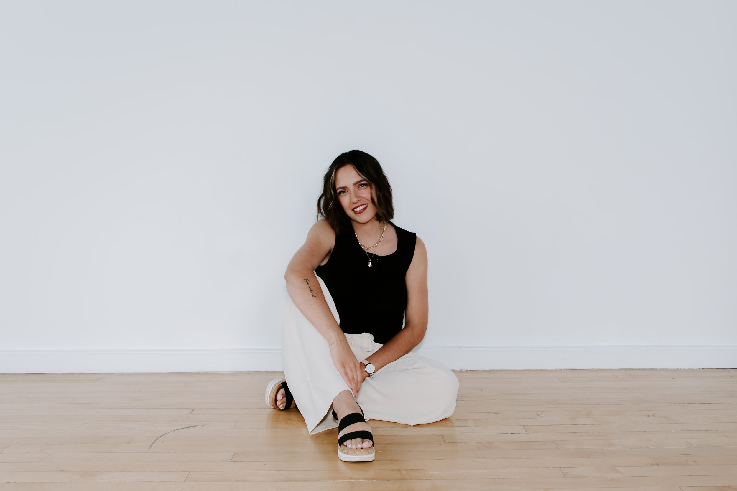 A woman with wavy dark hair sitting on a wooden floor against a plain white wall, smiling at the camera, wearing a black sleeveless top, white pants, and black sandals.
