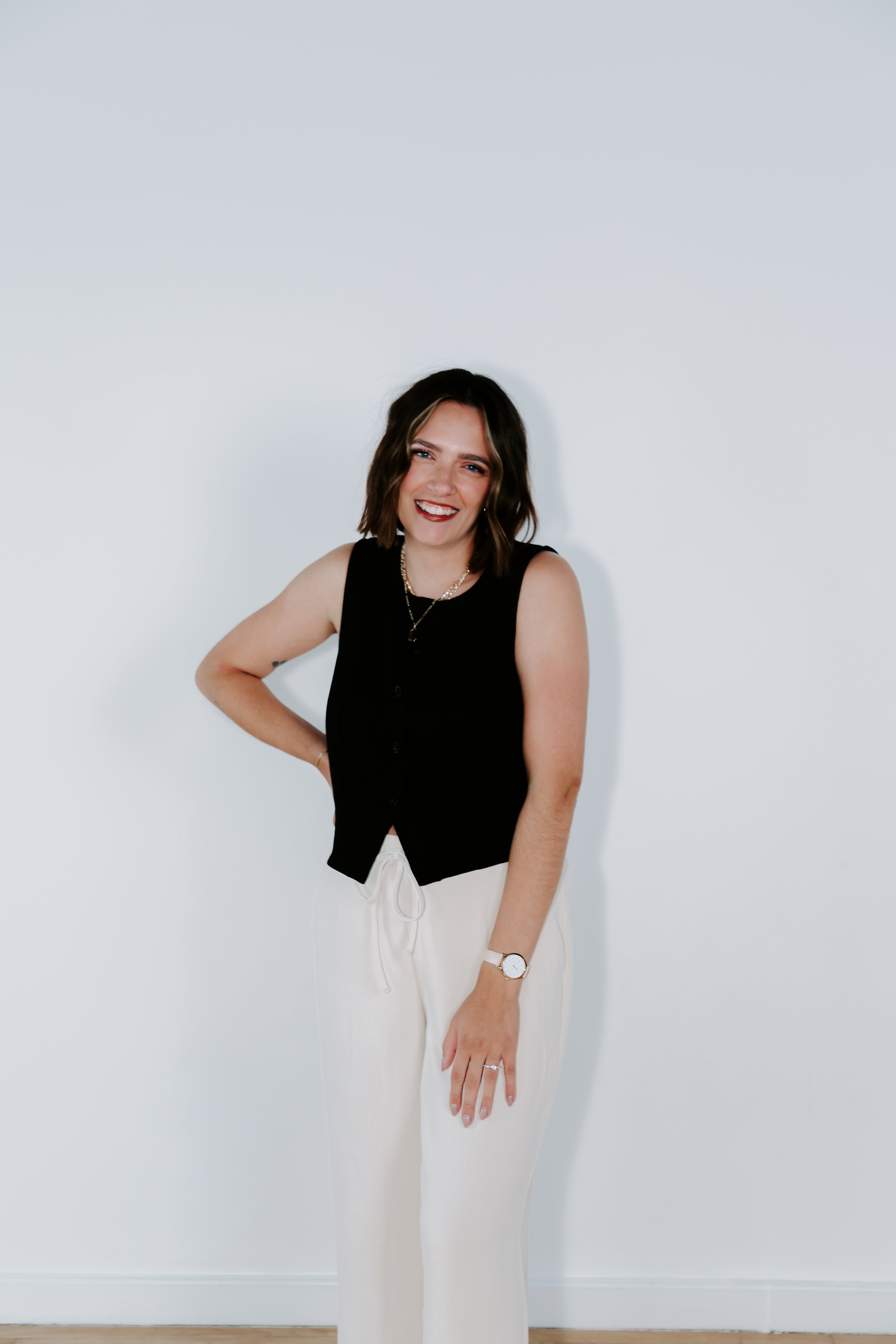 A woman with shoulder-length brown hair smiling, wearing a black sleeveless top, white pants, a watch, layered necklaces, and rings, standing against a plain white wall.