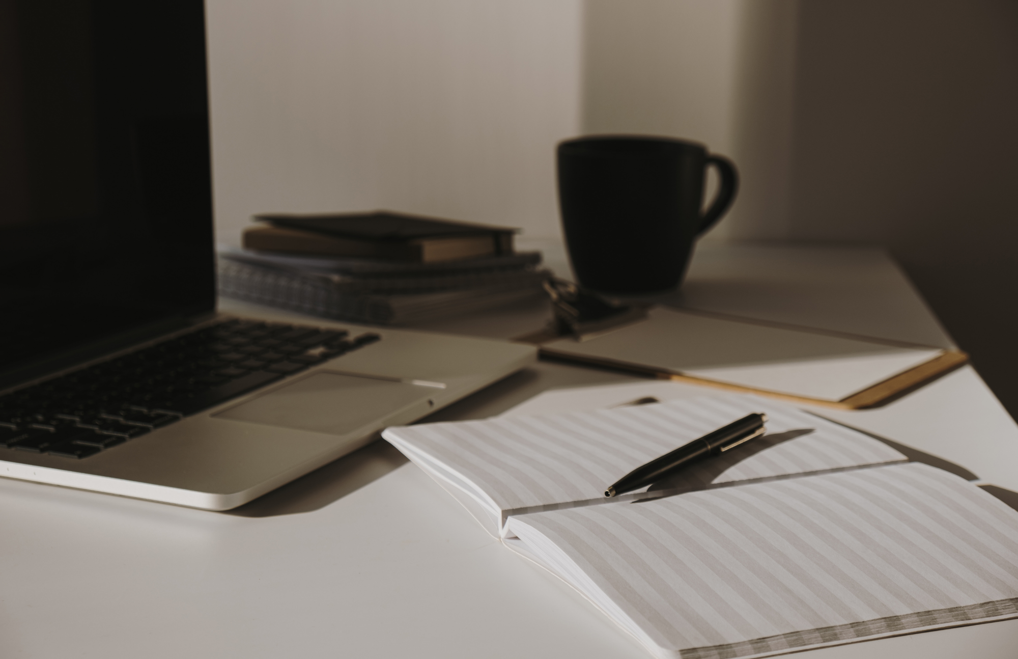 Desk with open notebook, pen, laptop, stacks of books, notebooks, a paperclip, and a dark coffee mug, with sunlight casting shadows.