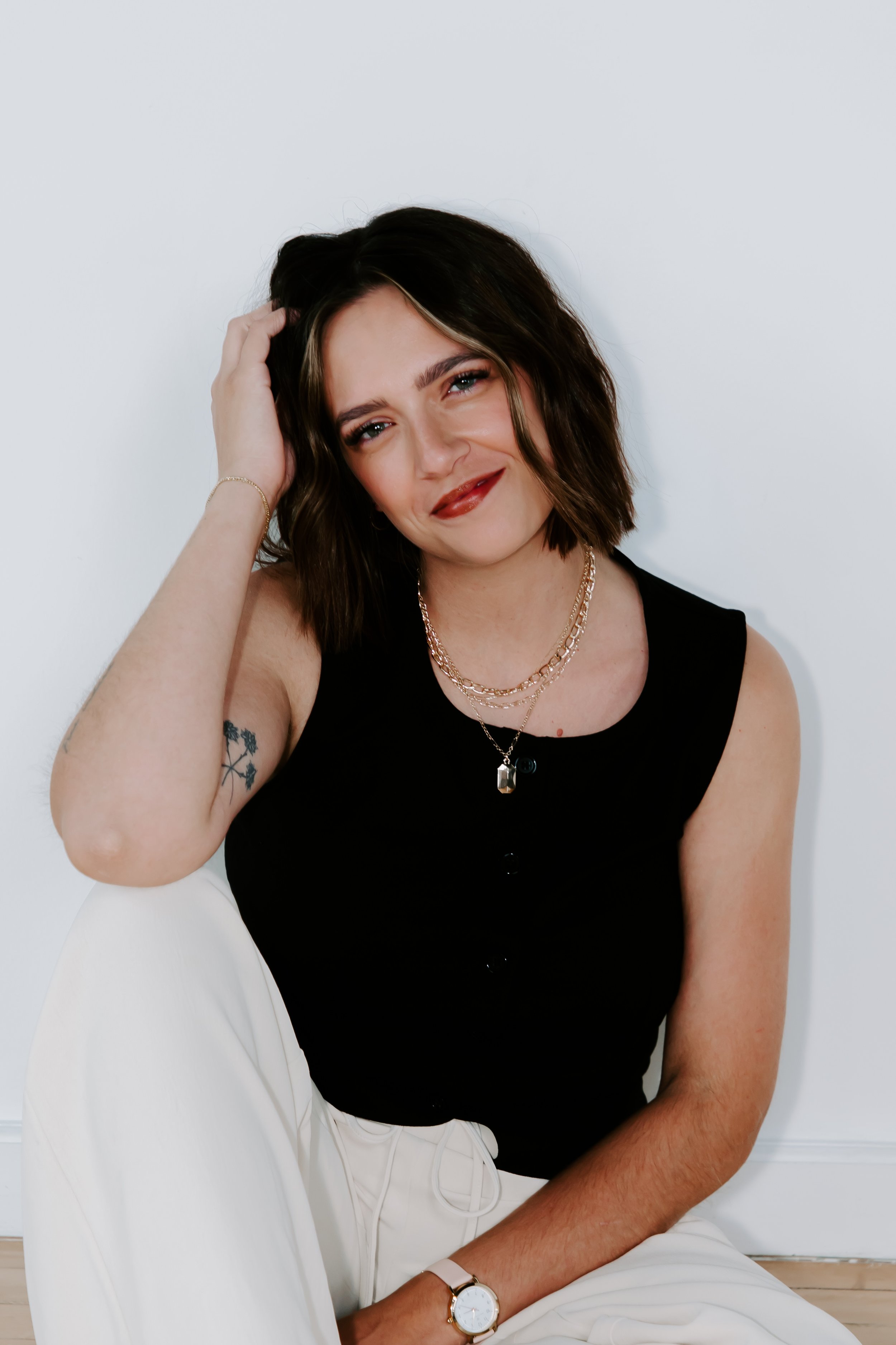 A woman with short dark hair, wearing a black sleeveless top, white pants, and gold jewelry, sitting against a white wall.