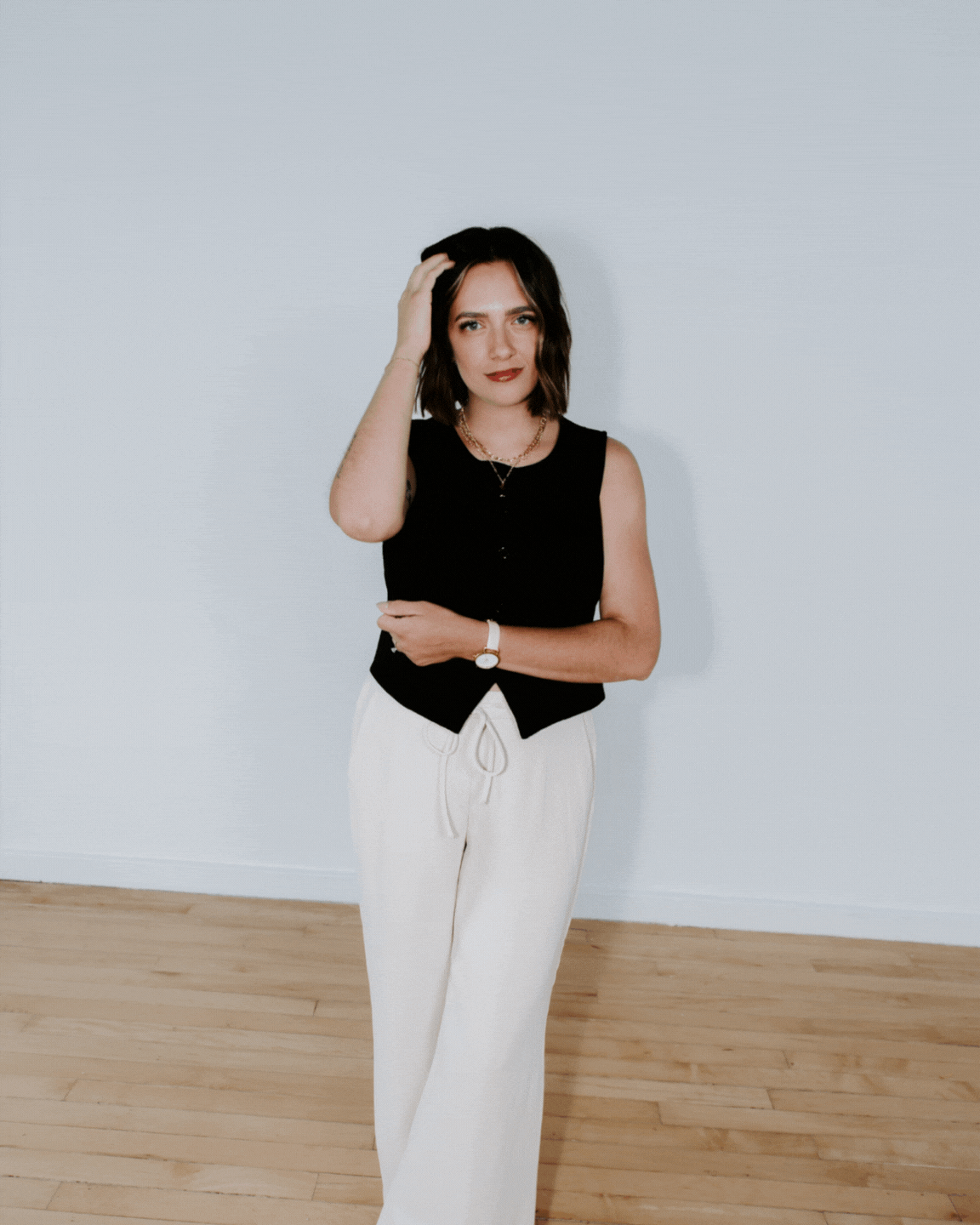 A young woman with dark wavy hair wearing a black sleeveless top and white drawstring pants, standing against a plain white wall with wooden flooring, touching her hair with a slight smile.