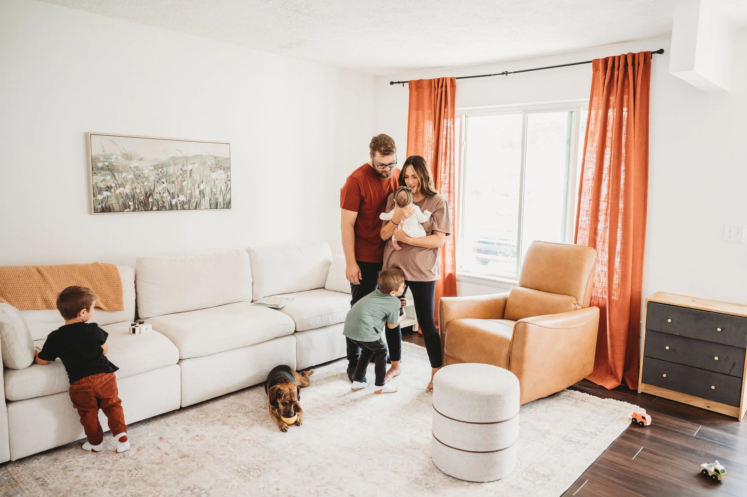 Family of five, two adults and three children, with a dog, in a living room with white walls, orange curtains, and a beige sofa.