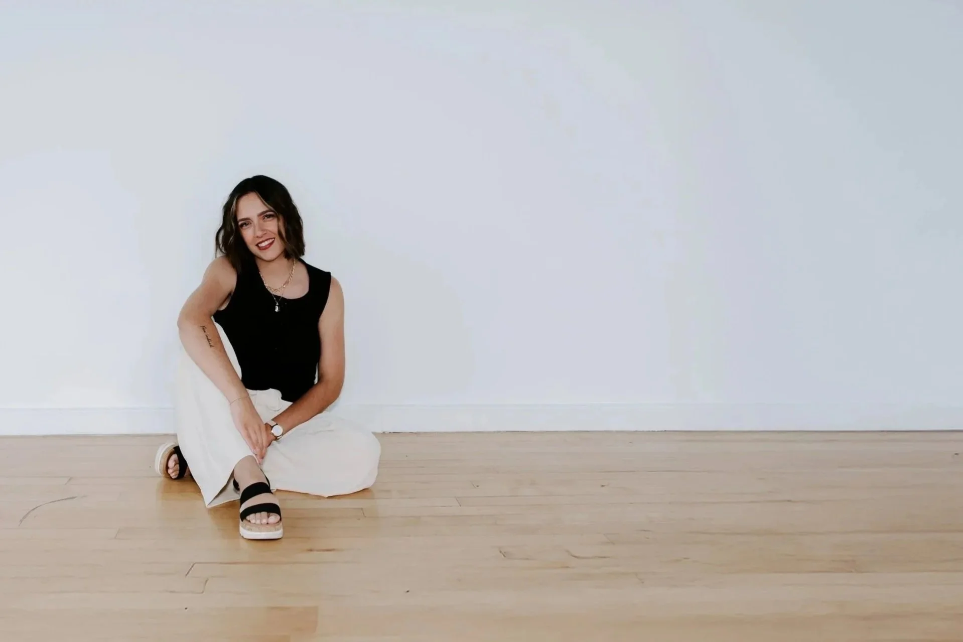 A woman with shoulder-length dark hair, wearing a black sleeveless top and white pants, is sitting on a light wooden floor against a plain white wall. She is smiling and looking at the camera.