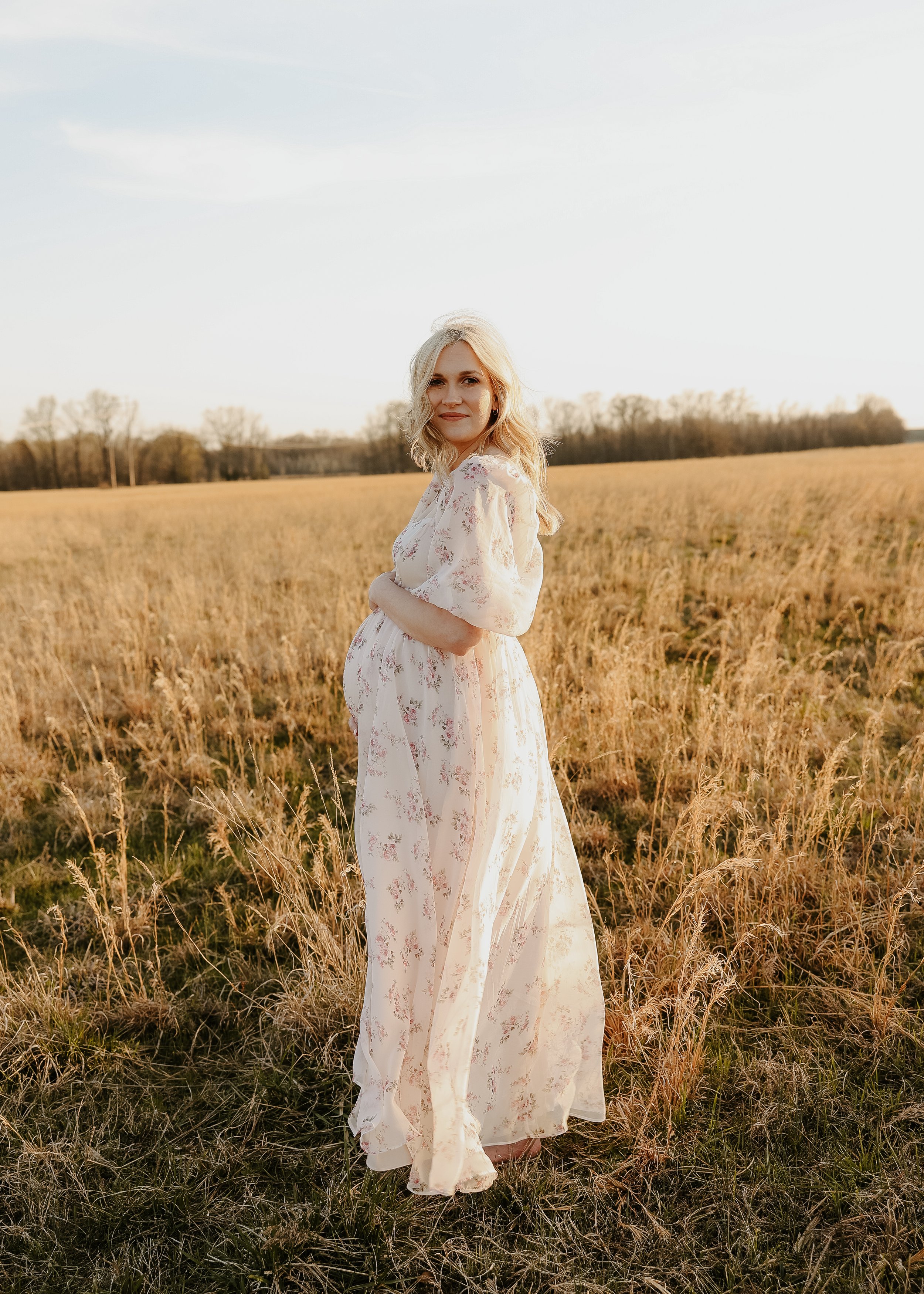 A blonde woman wearing a floral dress standing in a golden field during sunset