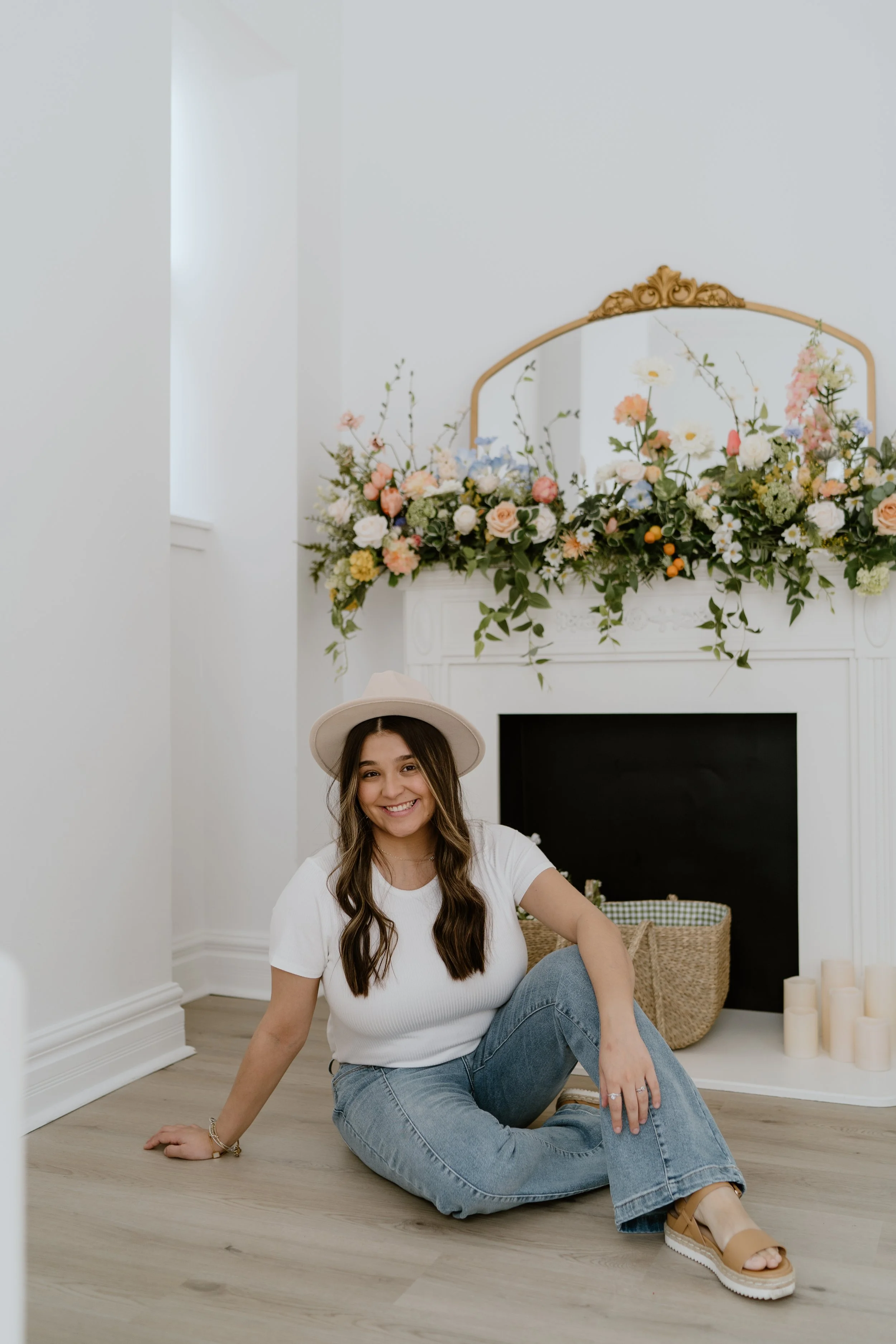A woman sitting on the floor near a white fireplace decorated with a large floral arrangement and candles, smiling at the camera.