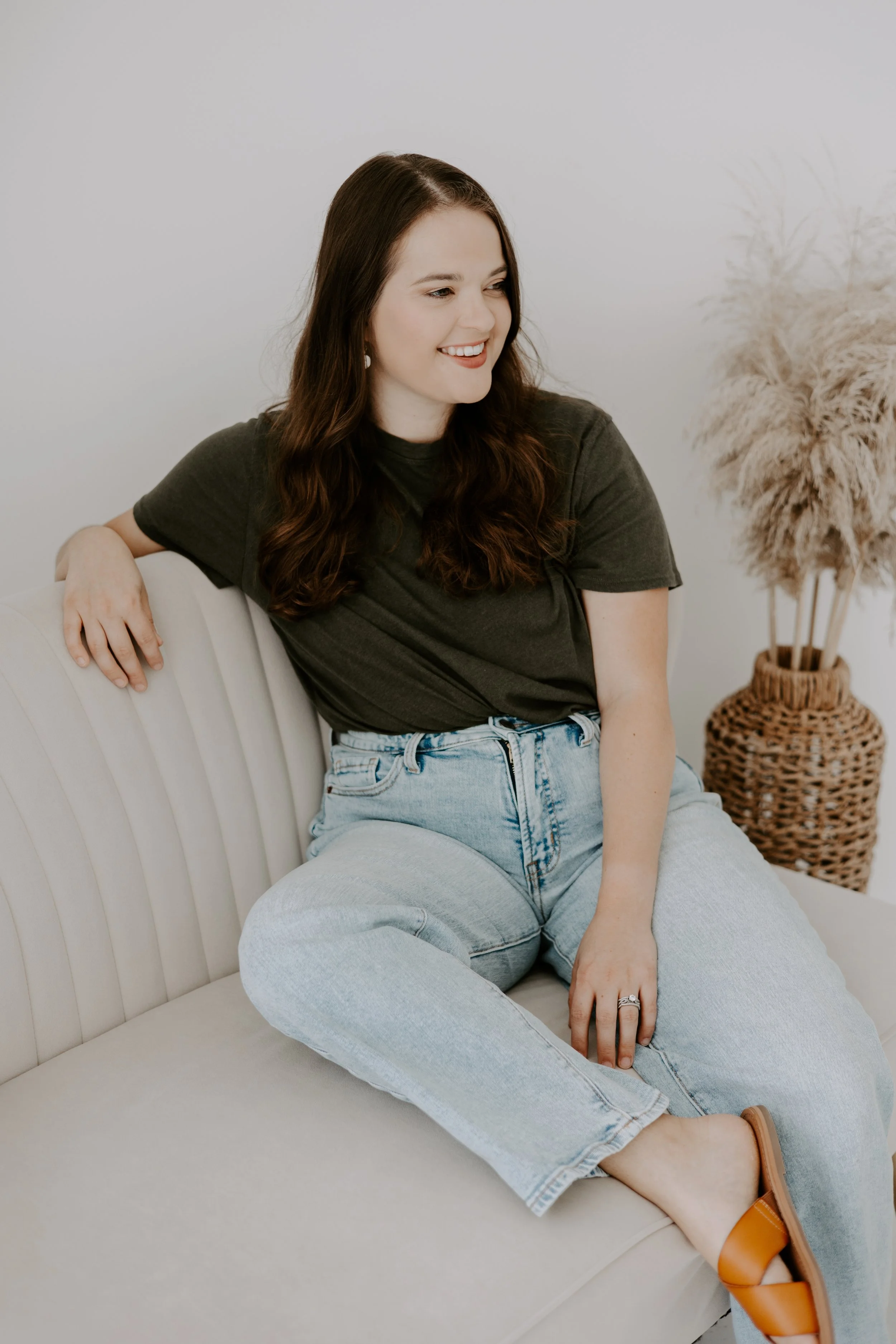 A woman with long, wavy brown hair sitting on a light-colored sofa, smiling and looking to her left, wearing a dark green t-shirt, light blue jeans, and tan slide sandals, with a large woven basket with dried plants next to her.