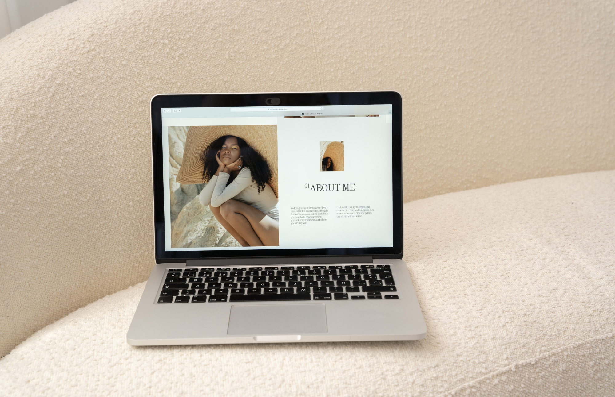 A silver laptop displaying a webpage with a photo of a woman with curly hair wearing a large straw hat, sitting outdoors on rocks, and an 'About Me' section on a beige textured background.