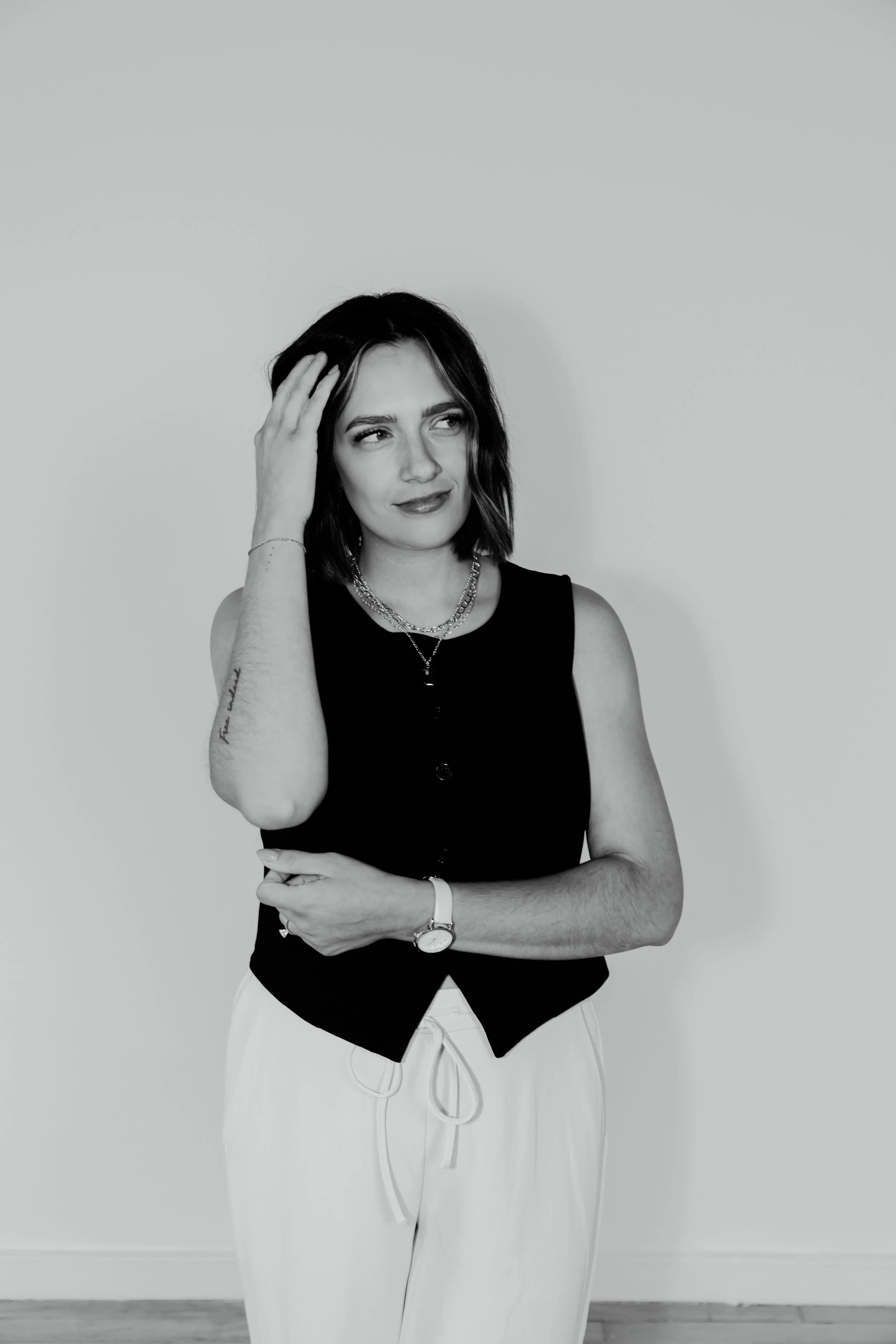 Black and white photo of a woman with shoulder-length dark hair, wearing a sleeveless black top, light-colored pants, and jewelry, standing against a plain background, looking to her left with a thoughtful expression.
