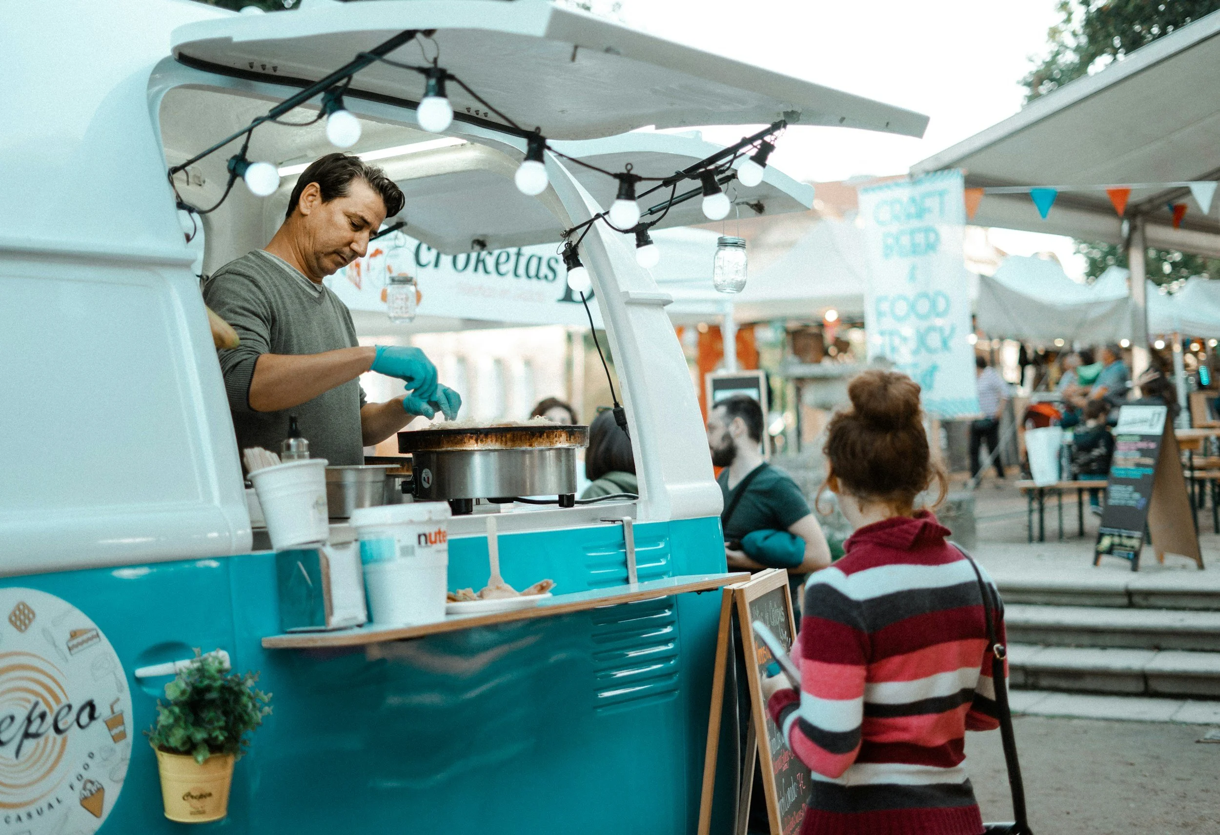 A food truck serving food at an outdoor market with a man cooking inside and a woman ordering from outside. The scene is decorated with string lights and signs for craft beer and food trucks.