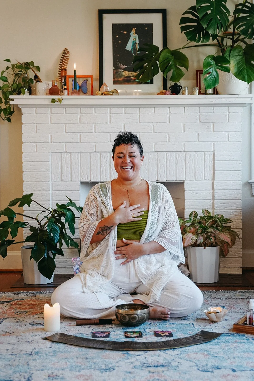 A woman sitting cross-legged on a rug in front of a white brick fireplace, smiling with her eyes closed, with her right hand over her heart and her left hand on her stomach, surrounded by candles, tarot cards, and plants.