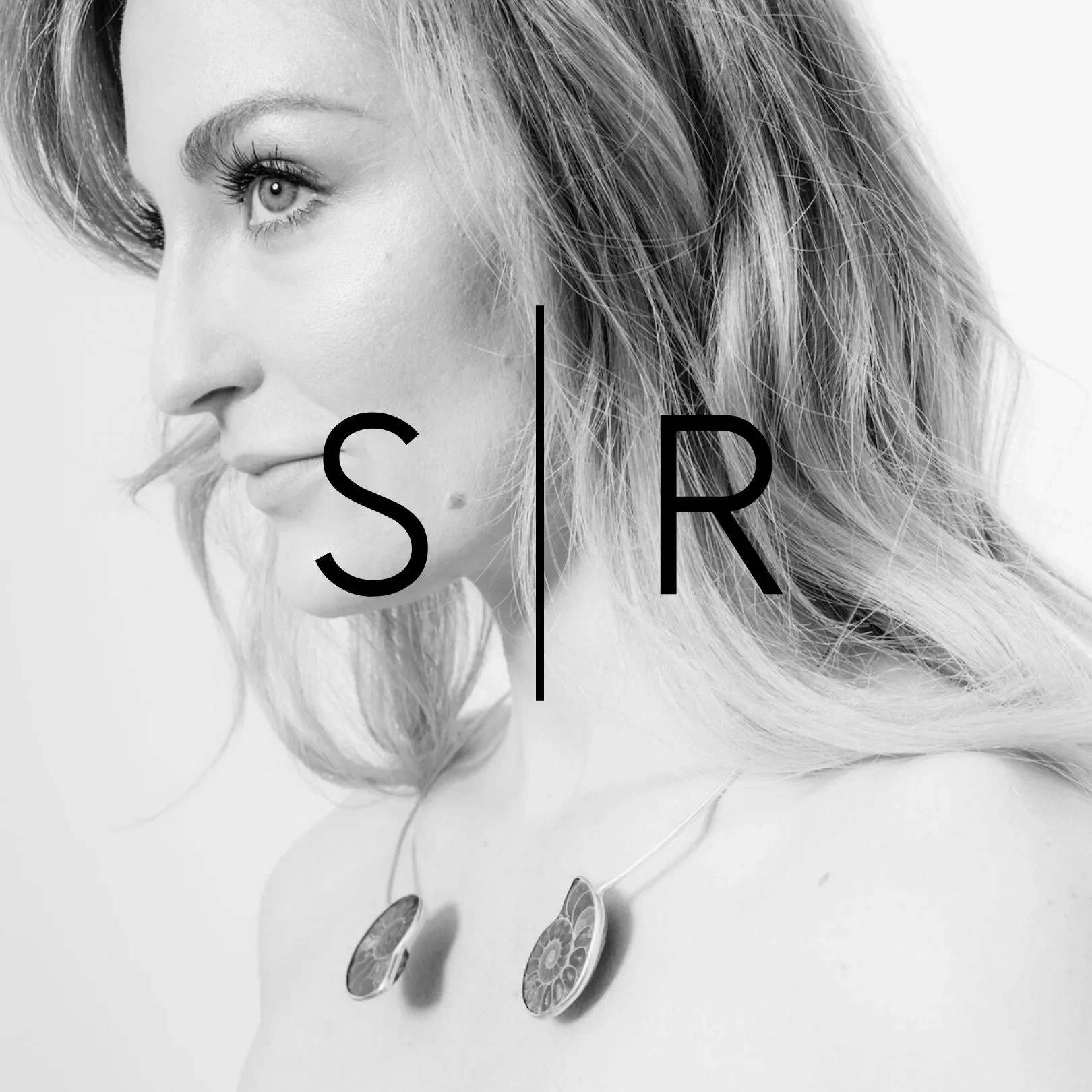 Black and white photo of a woman with long wavy hair, looking to the side. She is wearing a necklace with two circular pendants. The letters 'S' and 'R' are overlaid on the image separated by a vertical line.