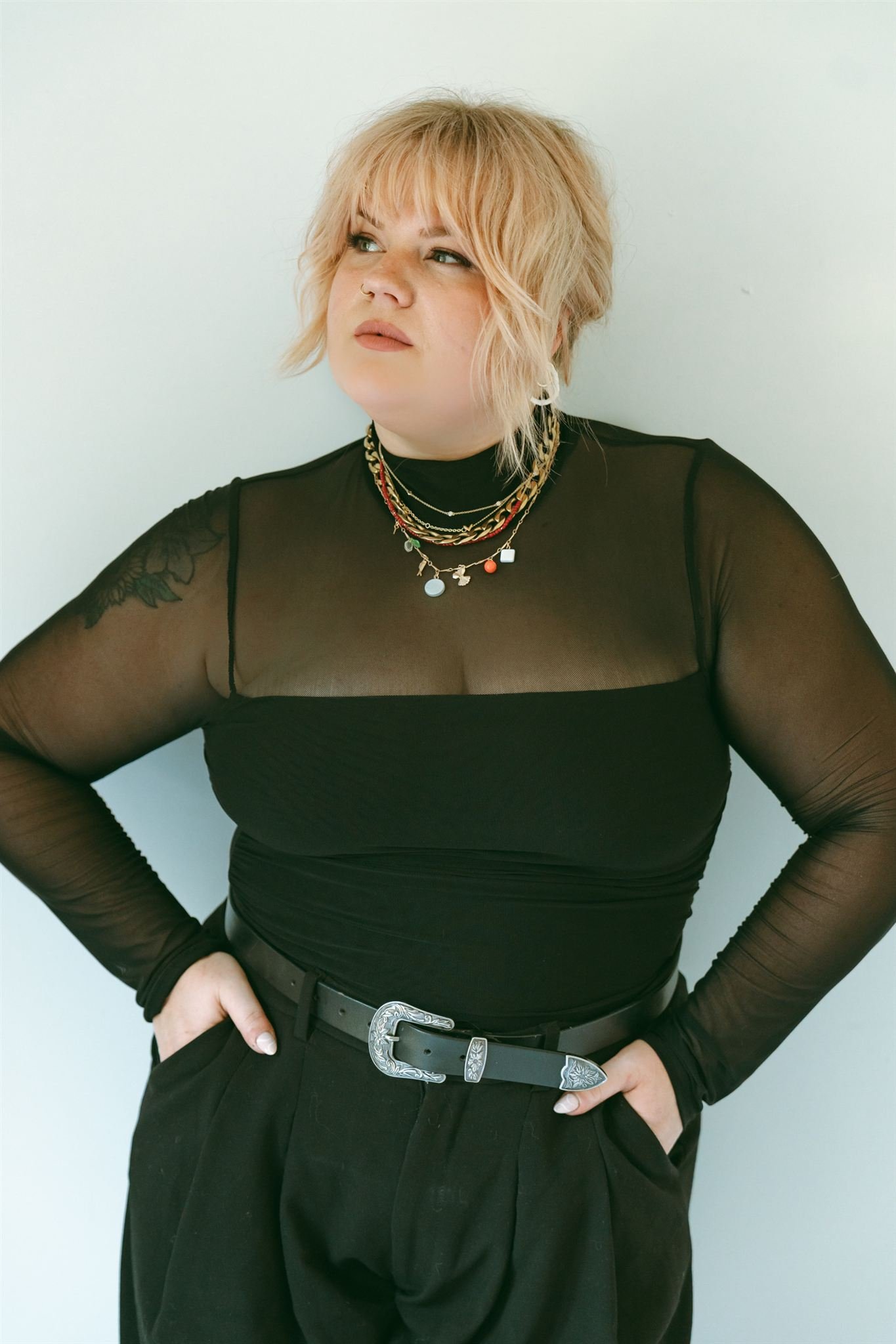 Woman with blonde, wavy hair wearing a black sheer top and layered necklaces, standing against a plain background.