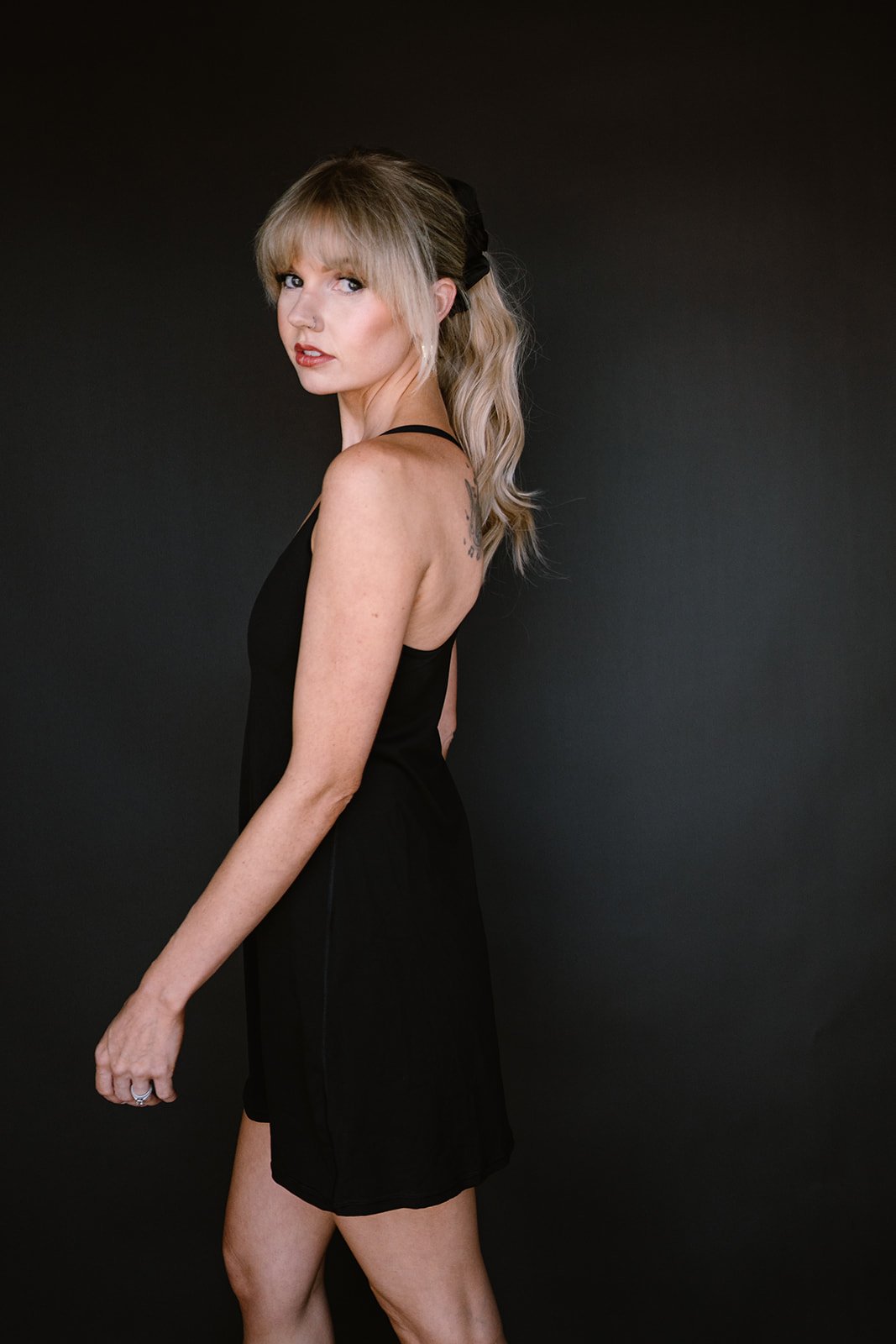 A woman with blonde hair in a ponytail, wearing a black sleeveless dress, standing against a dark background.