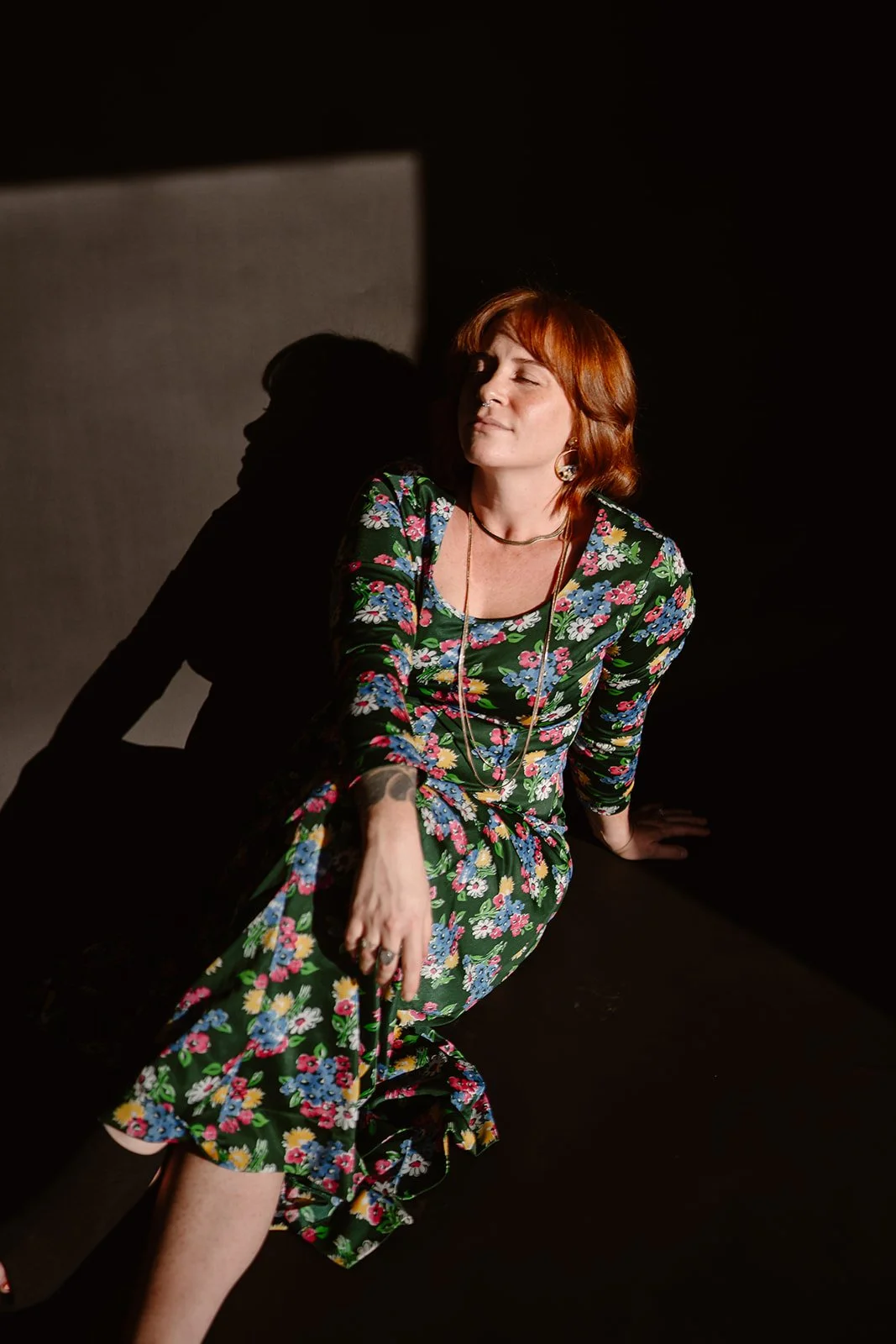 A woman with red hair and tattoos, wearing a floral dress, sitting on the floor with eyes closed against a dark background, illuminated by soft light.