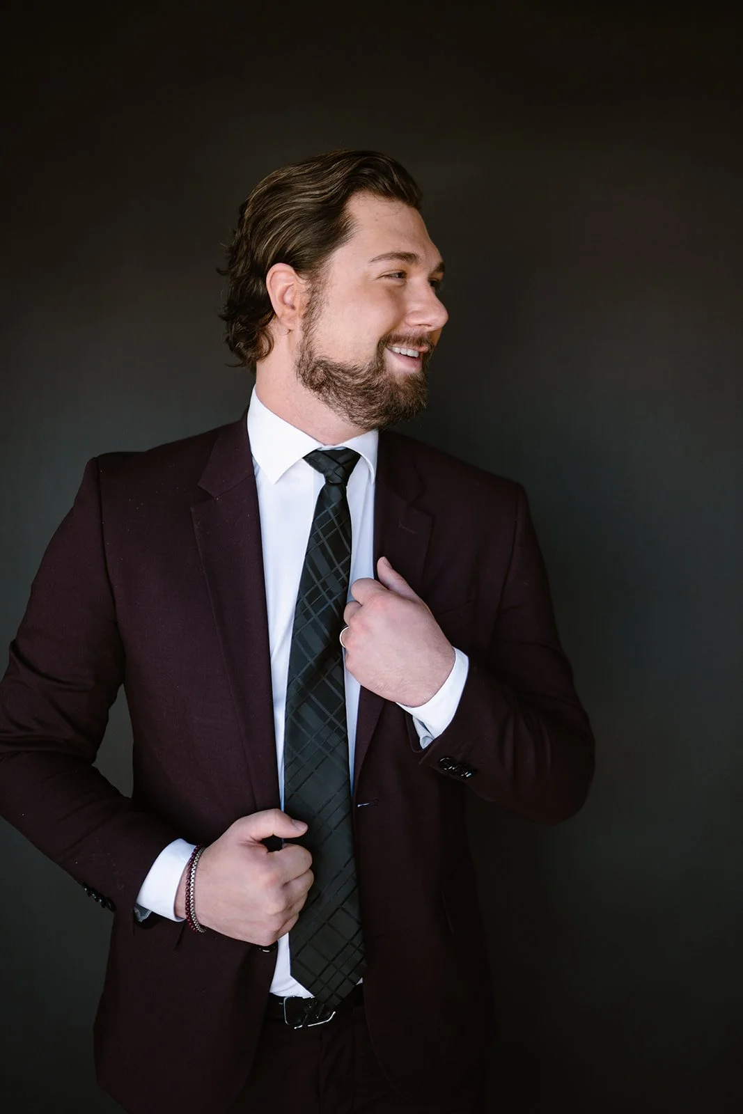 A man with brown hair and a beard wearing a dark suit, white shirt, and patterned tie, smiling and adjusting his jacket.