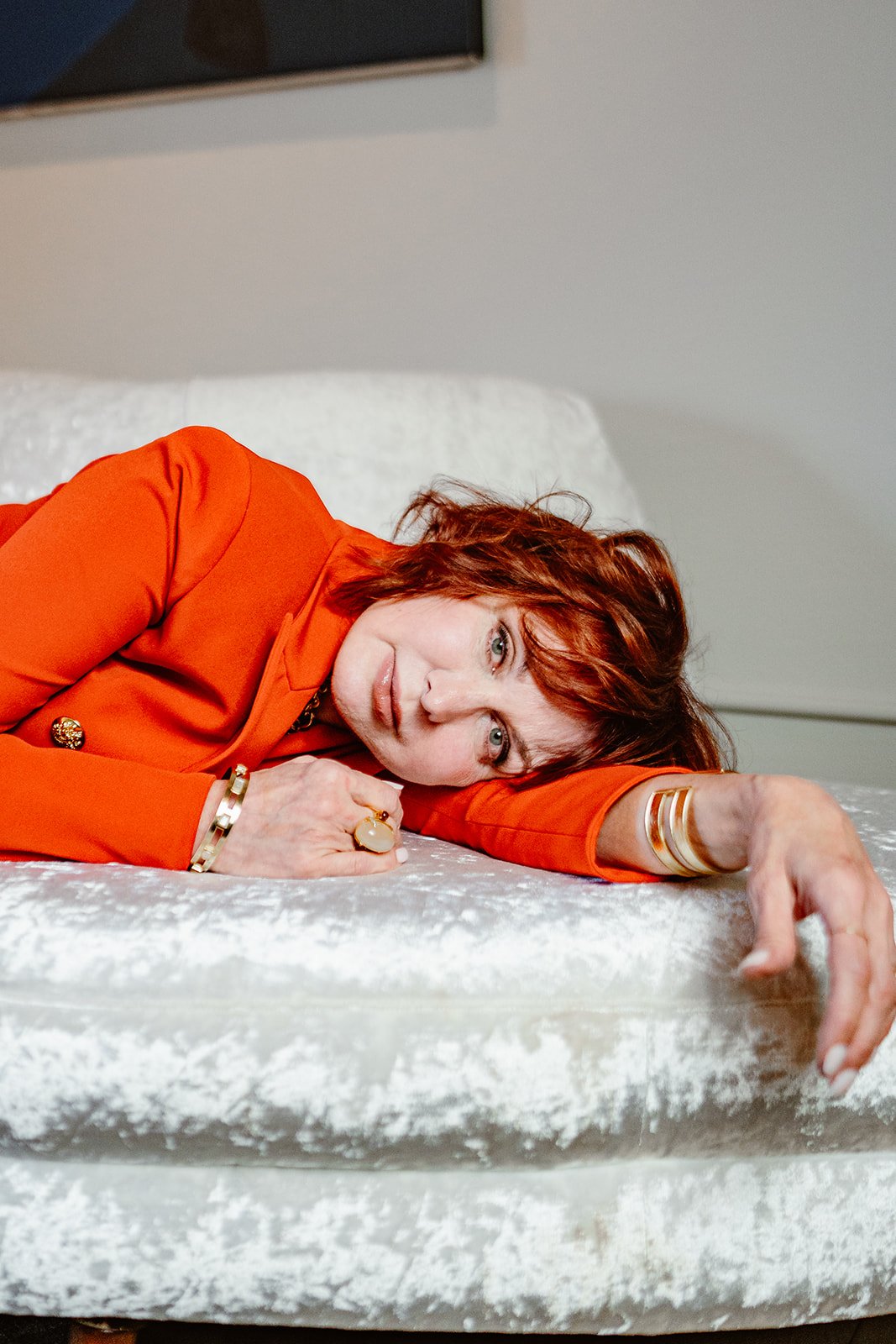 Woman with red hair wearing an orange blazer, lying on a white velvet bed, resting her head on her folded arm, looking at the camera.