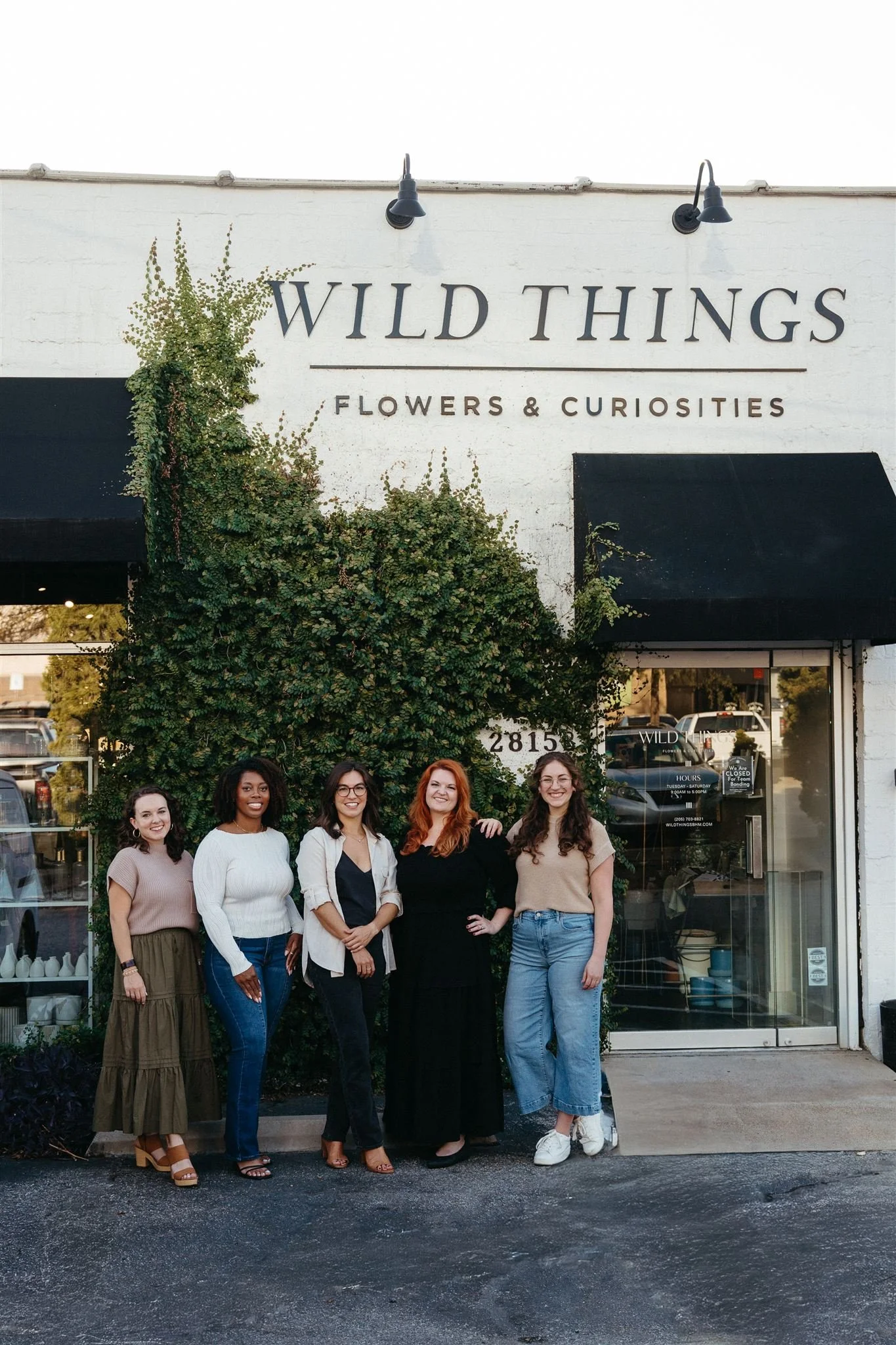 Five women standing outside a shop called 'Wild Things Flowers & Curiosities' with a large green bush and the shop's sign visible behind them.